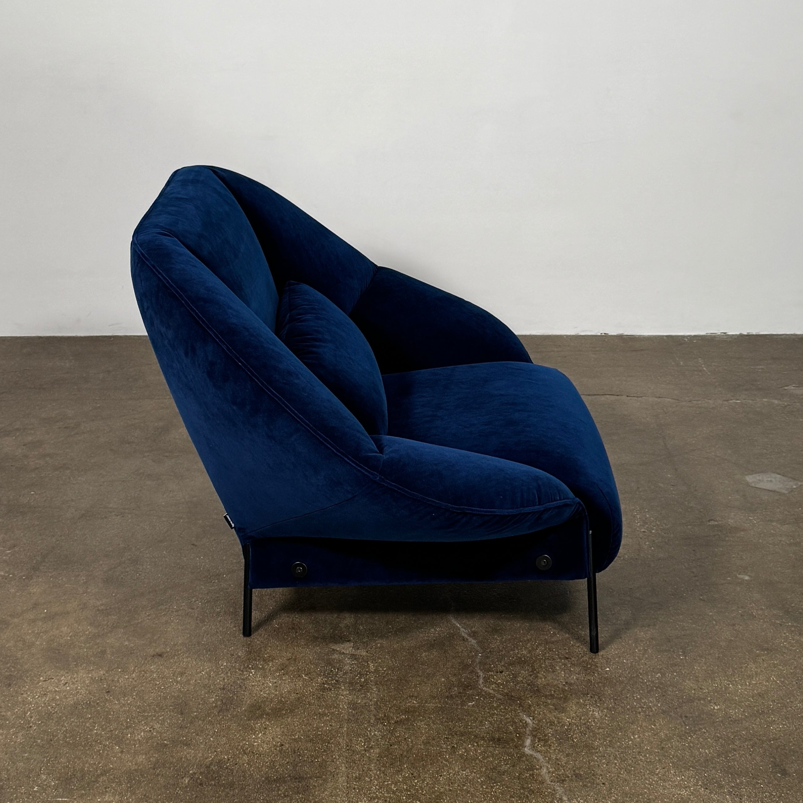 The Ligne Roset Paipai Armchair by Ligne Roset features a navy blue velvet finish, curved cocoon-like seat, matching rectangular cushion, slim black legs, and premium foam construction, displayed against a plain background.
