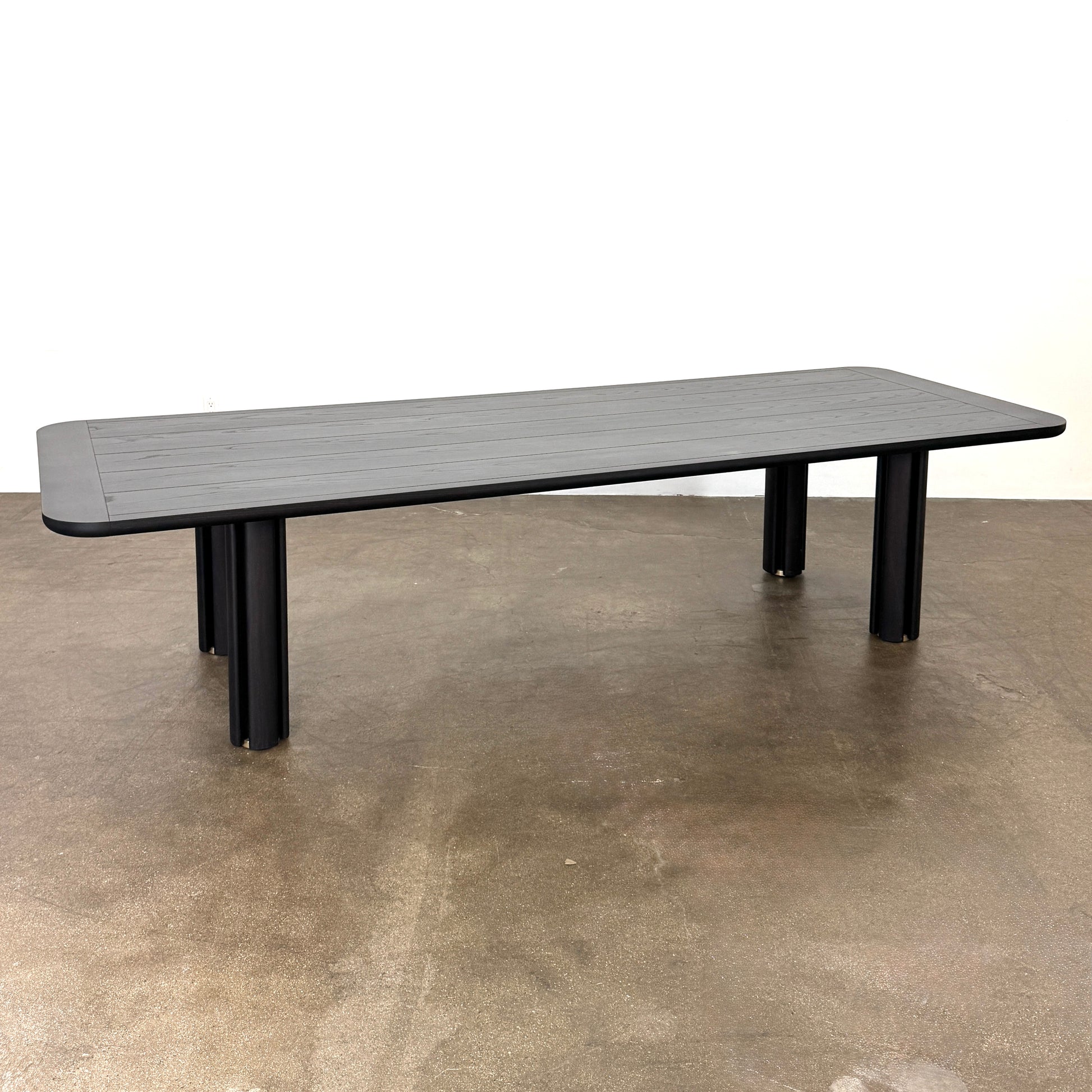 A rectangular black Porada Quadrifolgio Dining Table with rounded corners and four cylindrical legs stands on a brown floor against a plain white wall.