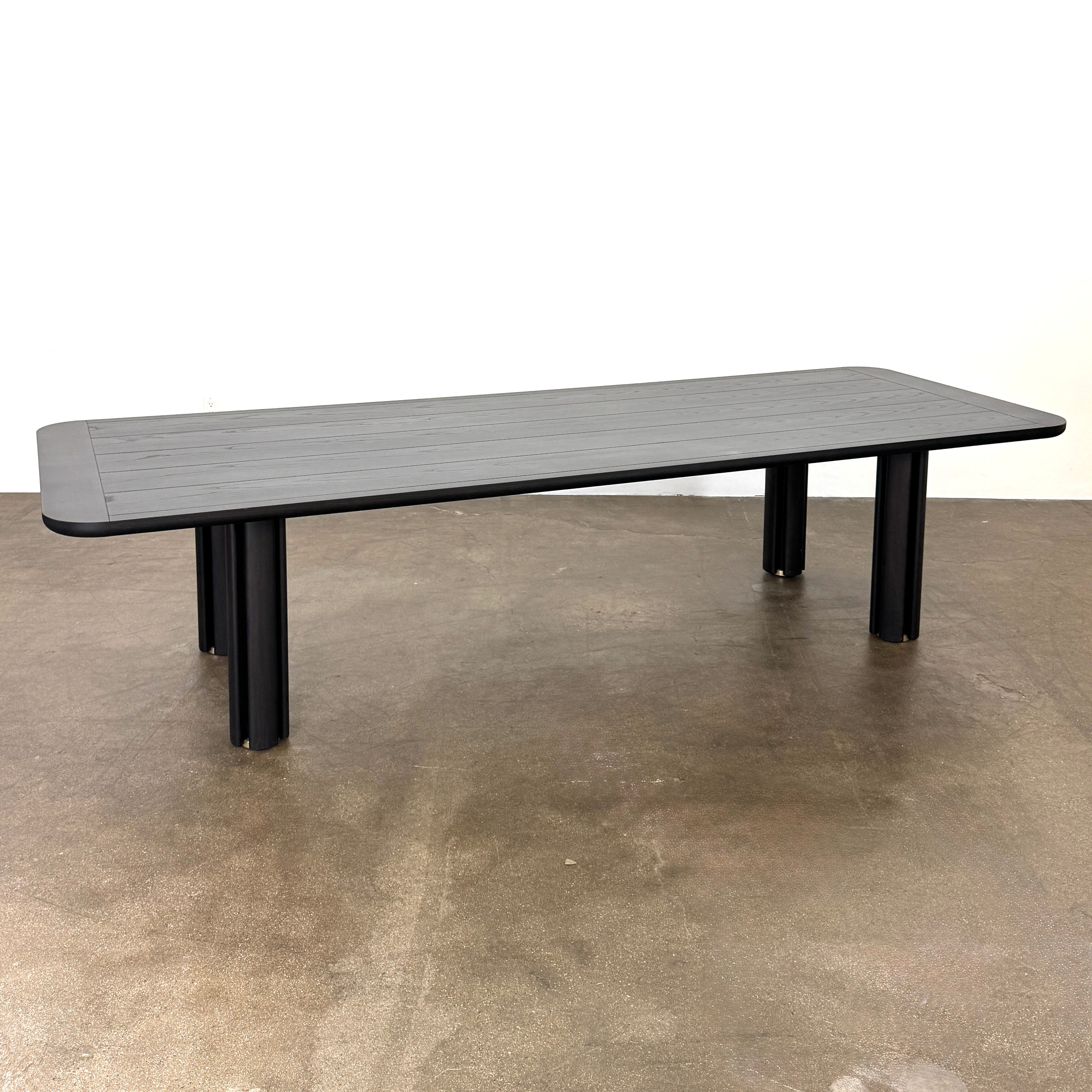 A rectangular black Porada Quadrifolgio Dining Table with rounded corners and four cylindrical legs stands on a brown floor against a plain white wall.