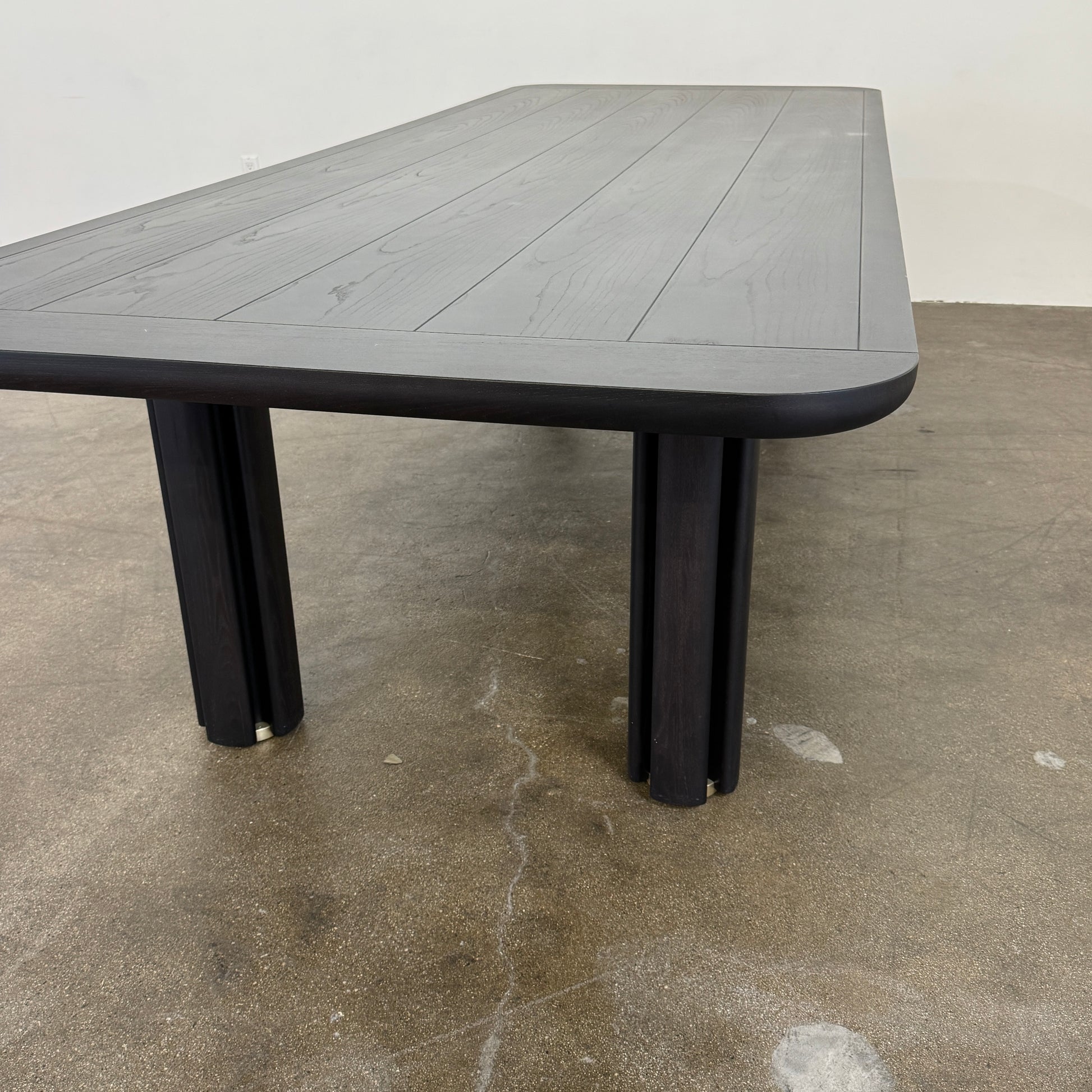A rectangular black Porada Quadrifolgio Dining Table with rounded corners and four cylindrical legs stands on a brown floor against a plain white wall.