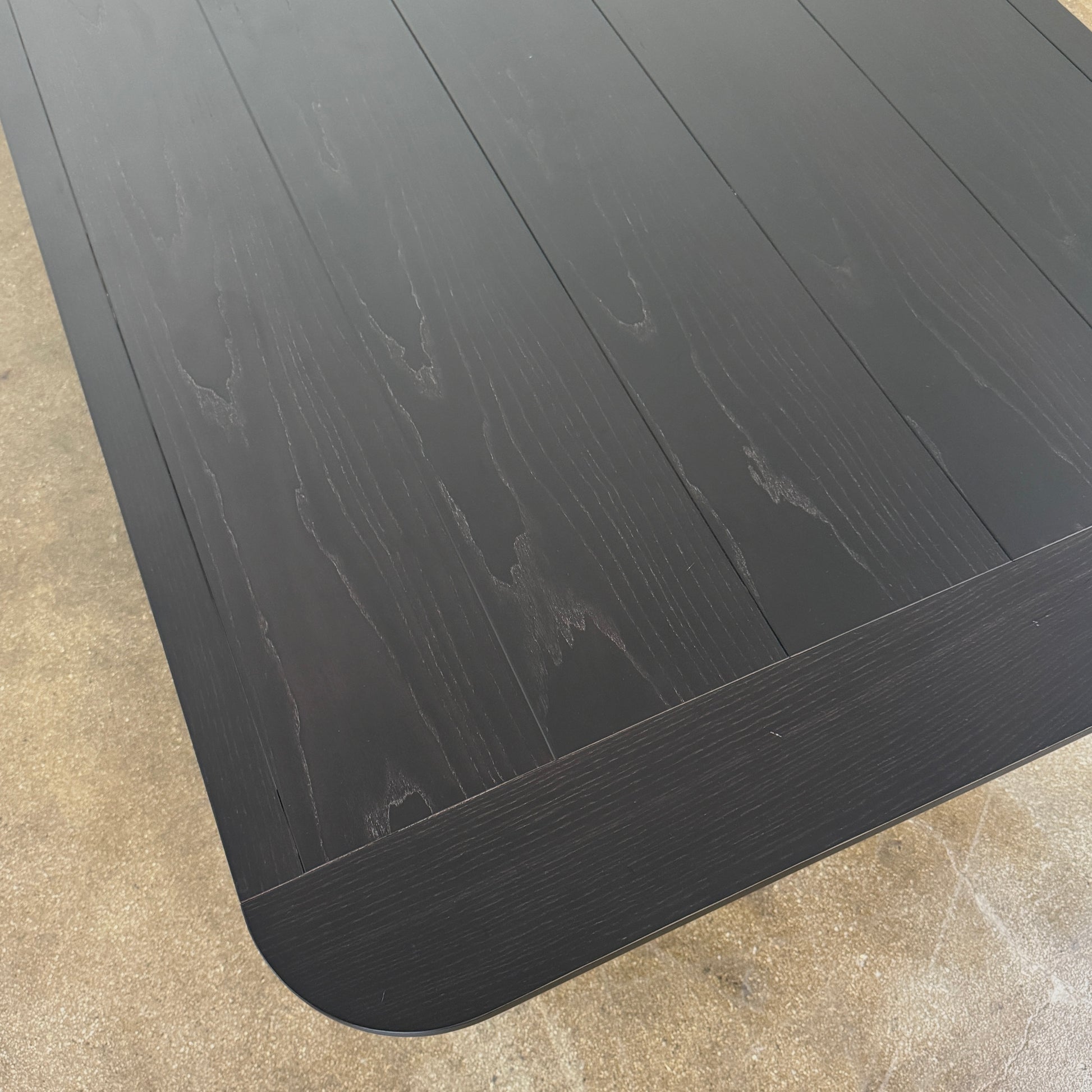 A rectangular black Porada Quadrifolgio Dining Table with rounded corners and four cylindrical legs stands on a brown floor against a plain white wall.