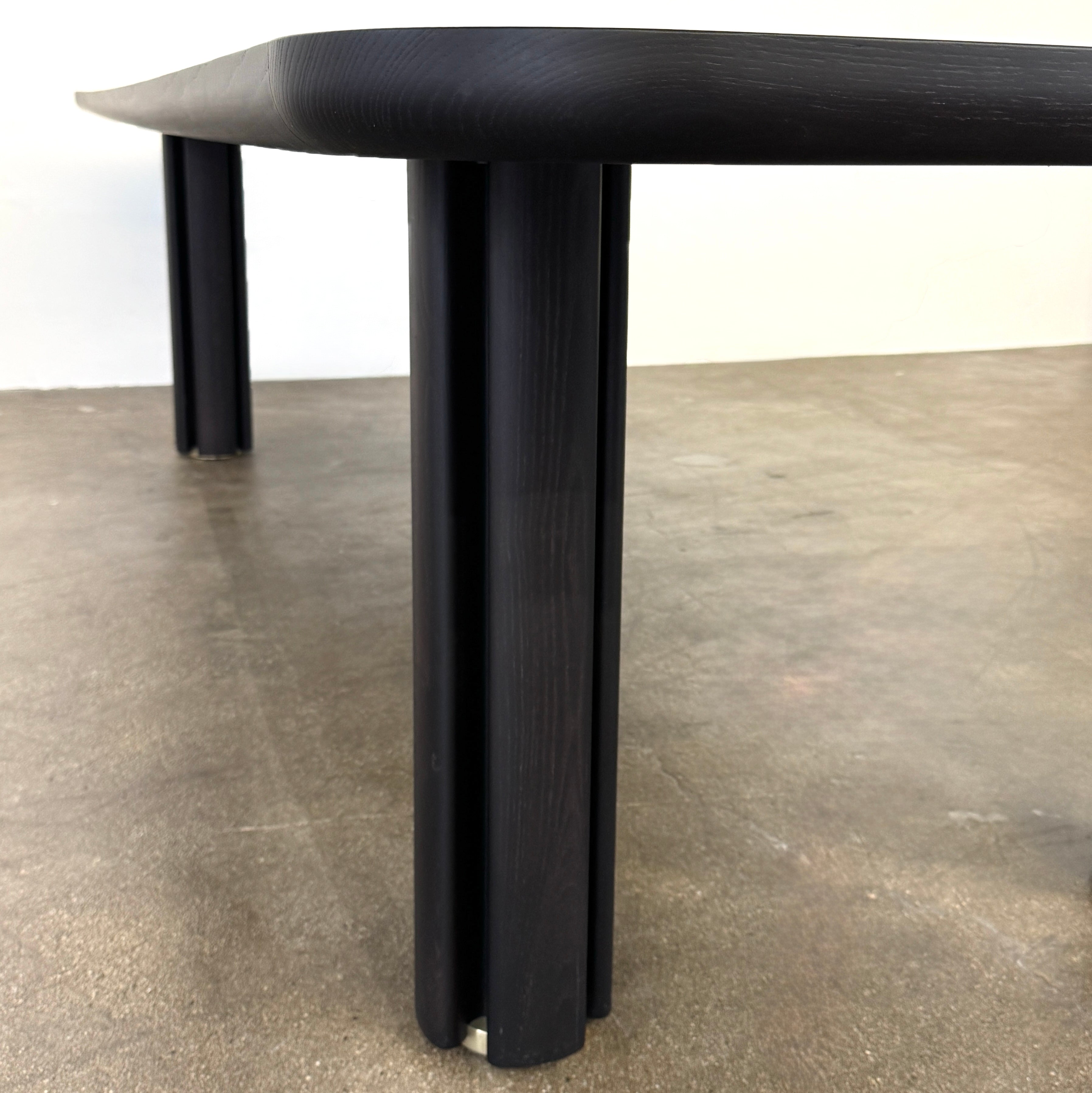 A rectangular black Porada Quadrifolgio Dining Table with rounded corners and four cylindrical legs stands on a brown floor against a plain white wall.