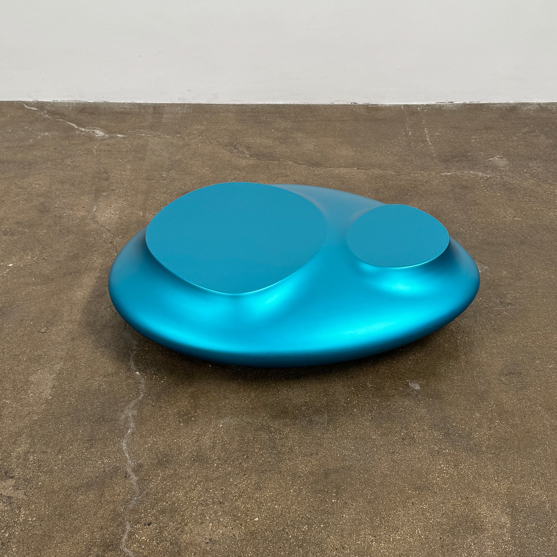 The Roche Bobois cute cut | XXM Cocktail Table by Roche Bobois is a smooth, metallic blue piece with rounded, uneven surfaces, displayed on a brown concrete floor against a white wall in contemporary style.