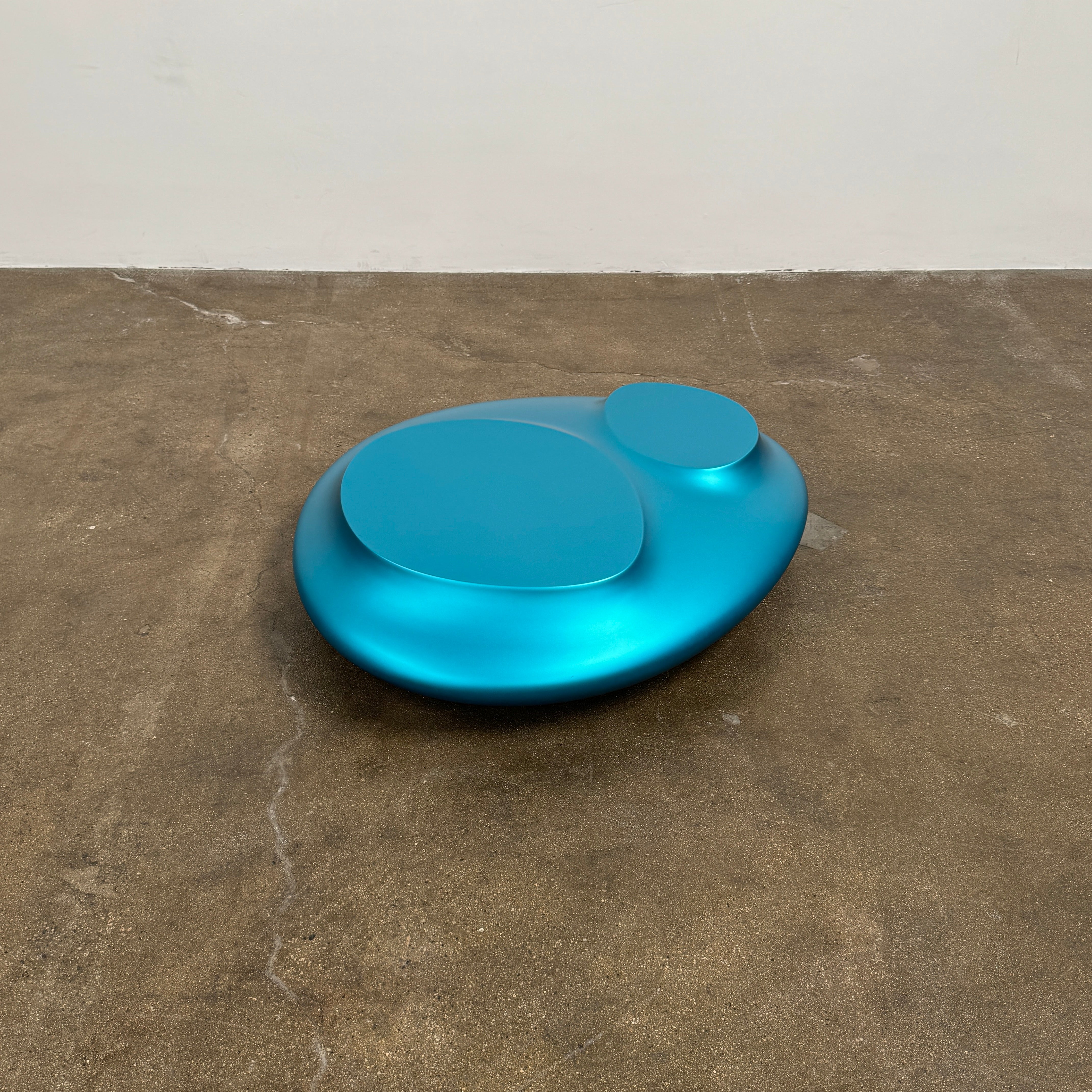 The Roche Bobois cute cut | XXM Cocktail Table by Roche Bobois is a smooth, metallic blue piece with rounded, uneven surfaces, displayed on a brown concrete floor against a white wall in contemporary style.