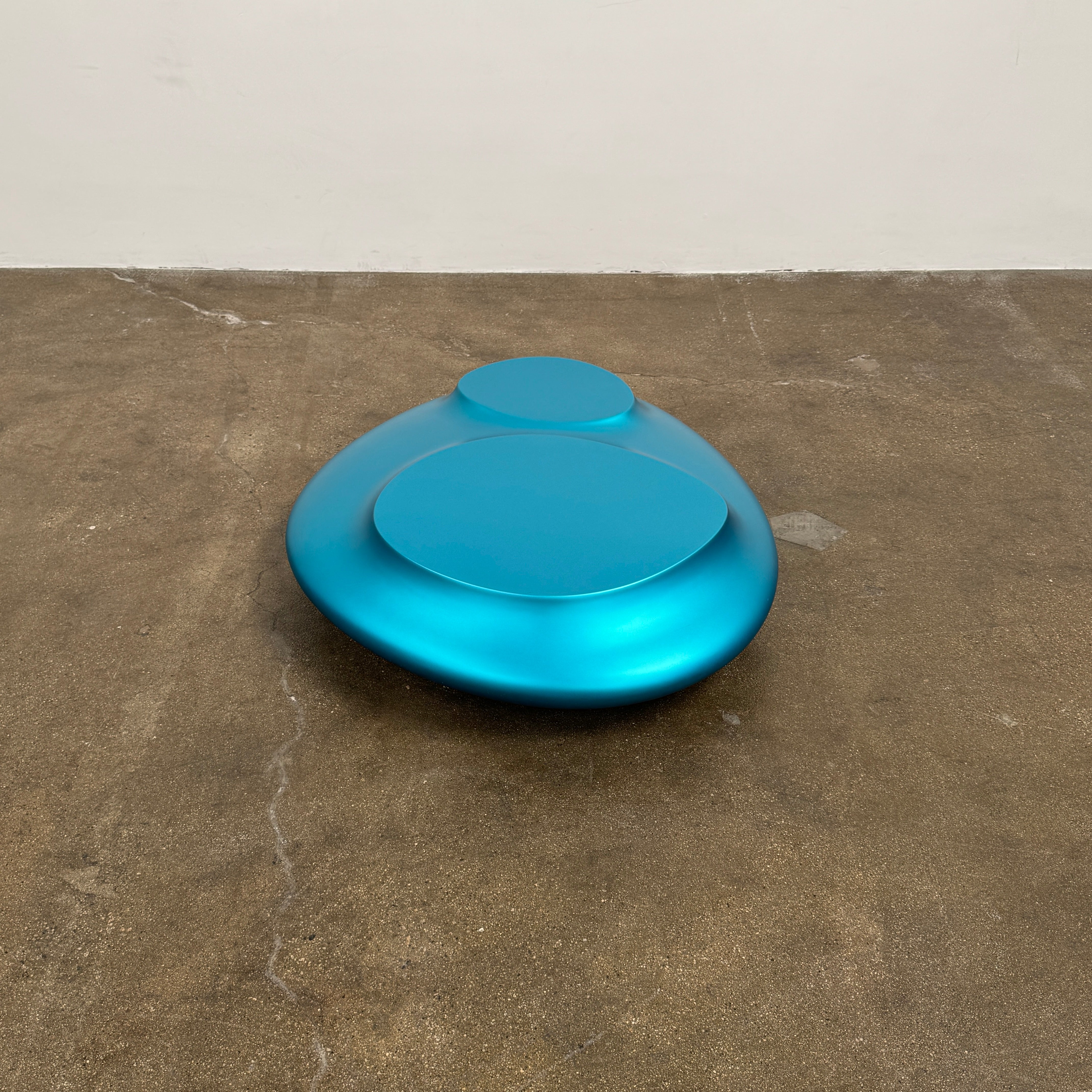 The Roche Bobois cute cut | XXM Cocktail Table by Roche Bobois is a smooth, metallic blue piece with rounded, uneven surfaces, displayed on a brown concrete floor against a white wall in contemporary style.