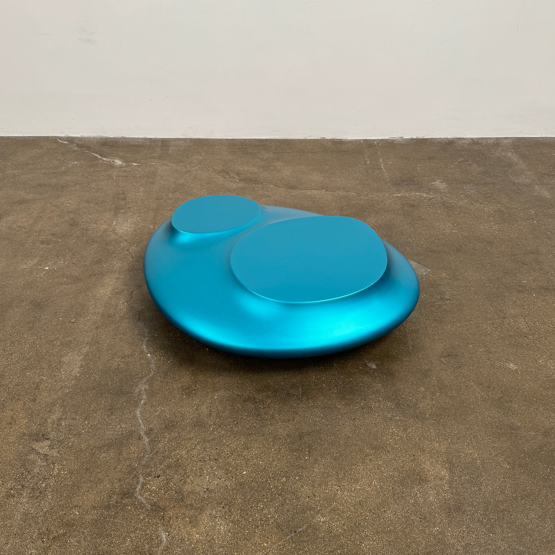The Roche Bobois cute cut | XXM Cocktail Table by Roche Bobois is a smooth, metallic blue piece with rounded, uneven surfaces, displayed on a brown concrete floor against a white wall in contemporary style.