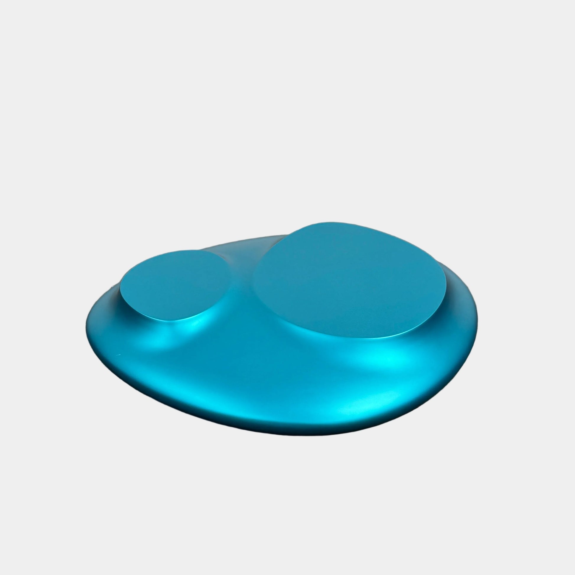The Roche Bobois cute cut | XXM Cocktail Table by Roche Bobois is a smooth, metallic blue piece with rounded, uneven surfaces, displayed on a brown concrete floor against a white wall in contemporary style.