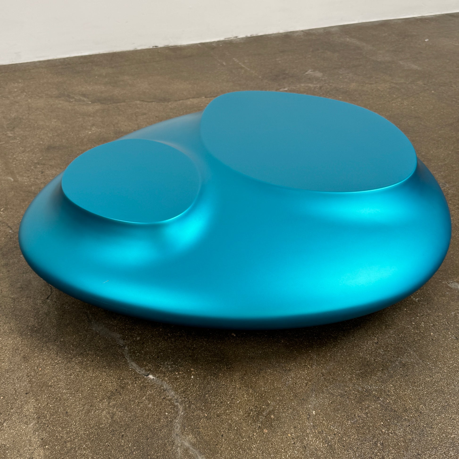 The Roche Bobois cute cut | XXM Cocktail Table by Roche Bobois is a smooth, metallic blue piece with rounded, uneven surfaces, displayed on a brown concrete floor against a white wall in contemporary style.