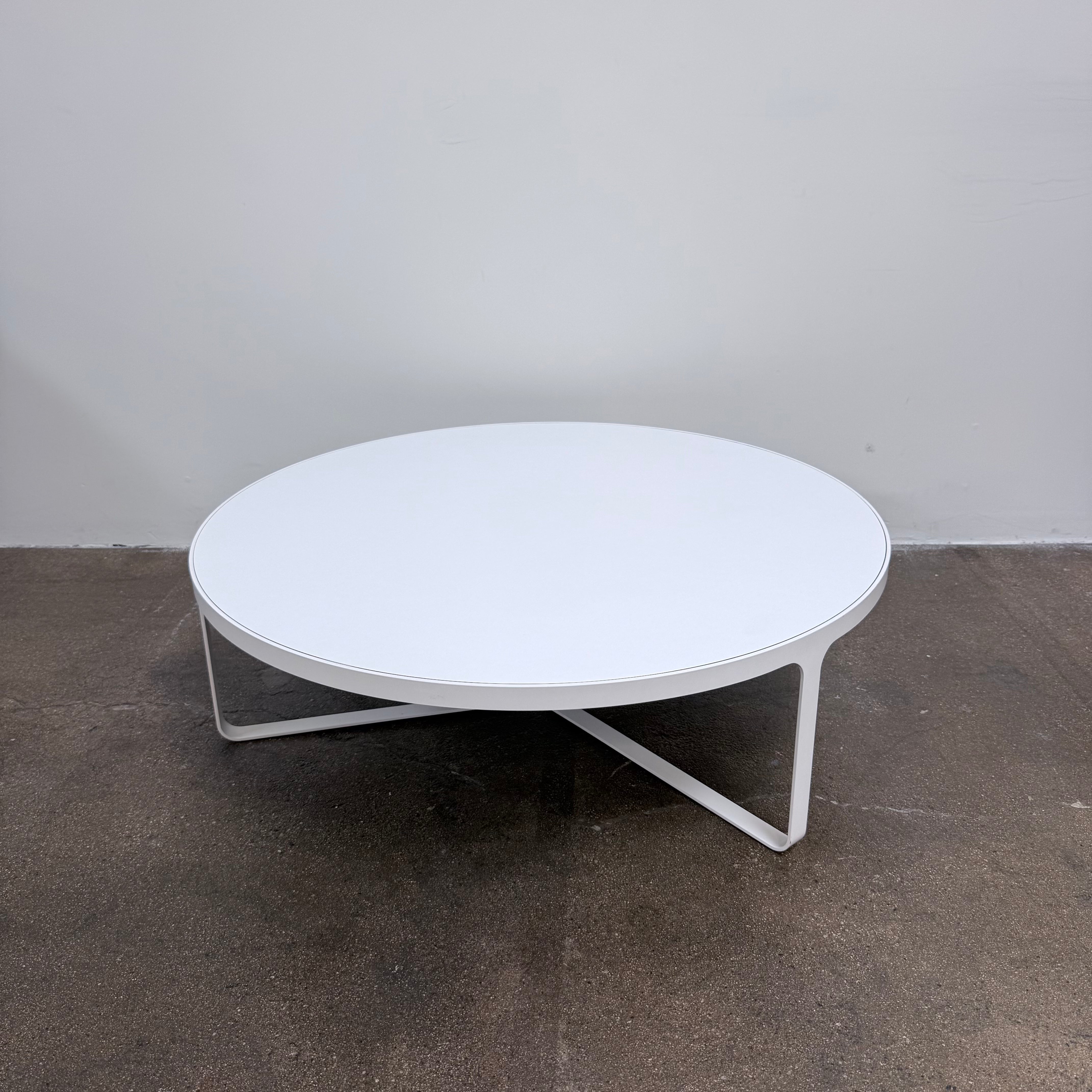 A round Tacchini Cage Coffee Table, White by Tacchini with thin metal legs stands on a plain gray concrete floor against a blank wall.