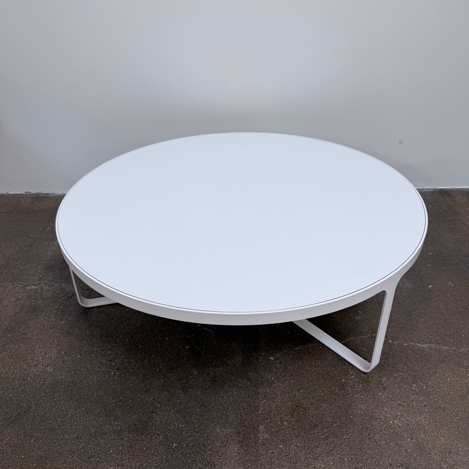 A round Tacchini Cage Coffee Table, White by Tacchini with thin metal legs stands on a plain gray concrete floor against a blank wall.
