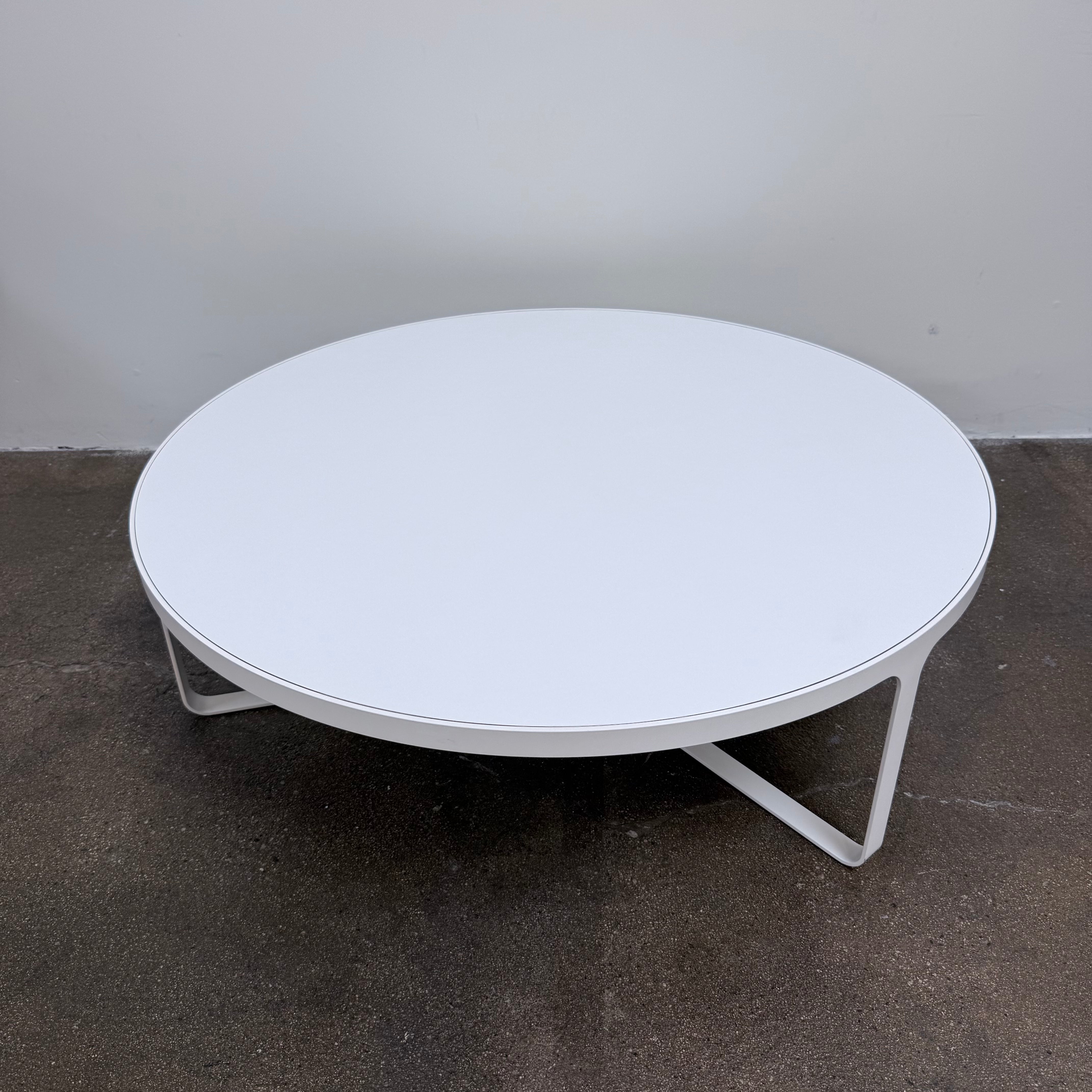 A round Tacchini Cage Coffee Table, White by Tacchini with thin metal legs stands on a plain gray concrete floor against a blank wall.