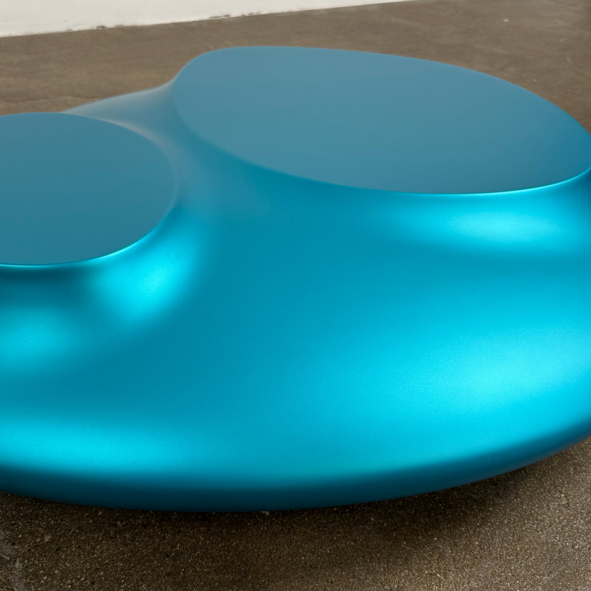 The Roche Bobois cute cut | XXM Cocktail Table by Roche Bobois is a smooth, metallic blue piece with rounded, uneven surfaces, displayed on a brown concrete floor against a white wall in contemporary style.