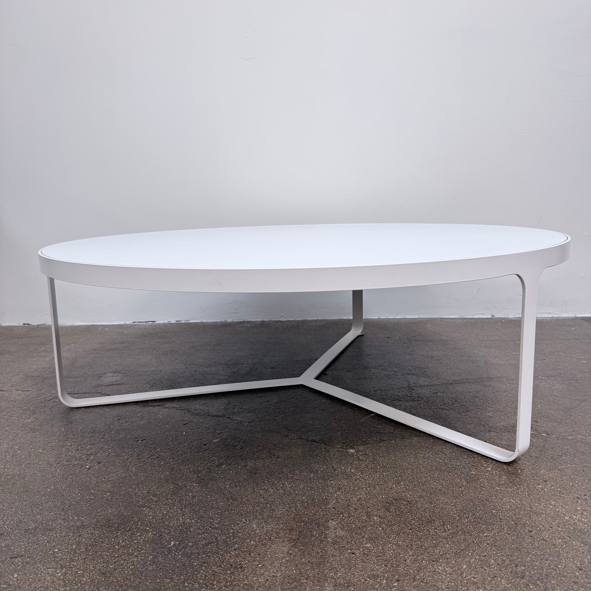 A round Tacchini Cage Coffee Table, White by Tacchini with thin metal legs stands on a plain gray concrete floor against a blank wall.
