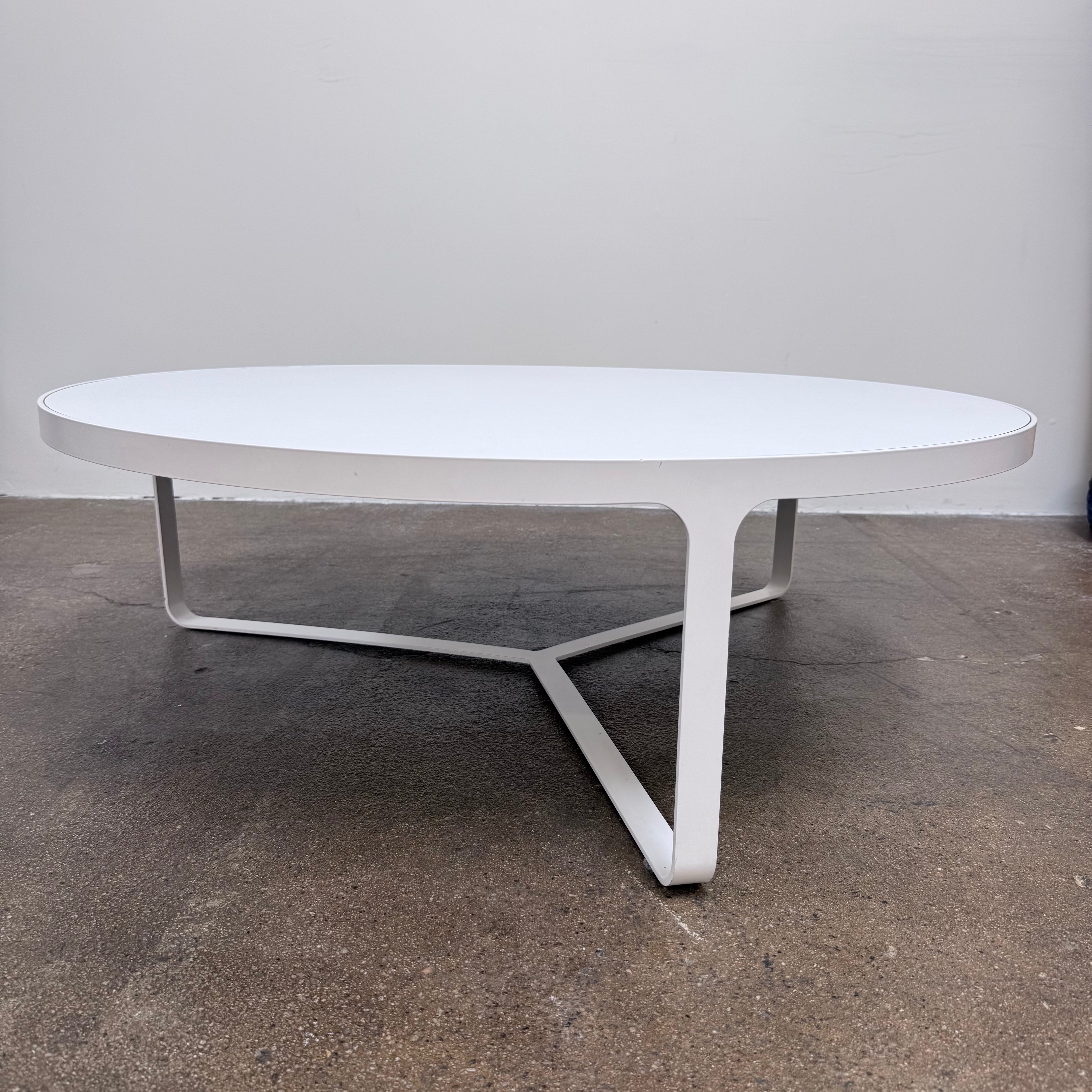 A round Tacchini Cage Coffee Table, White by Tacchini with thin metal legs stands on a plain gray concrete floor against a blank wall.