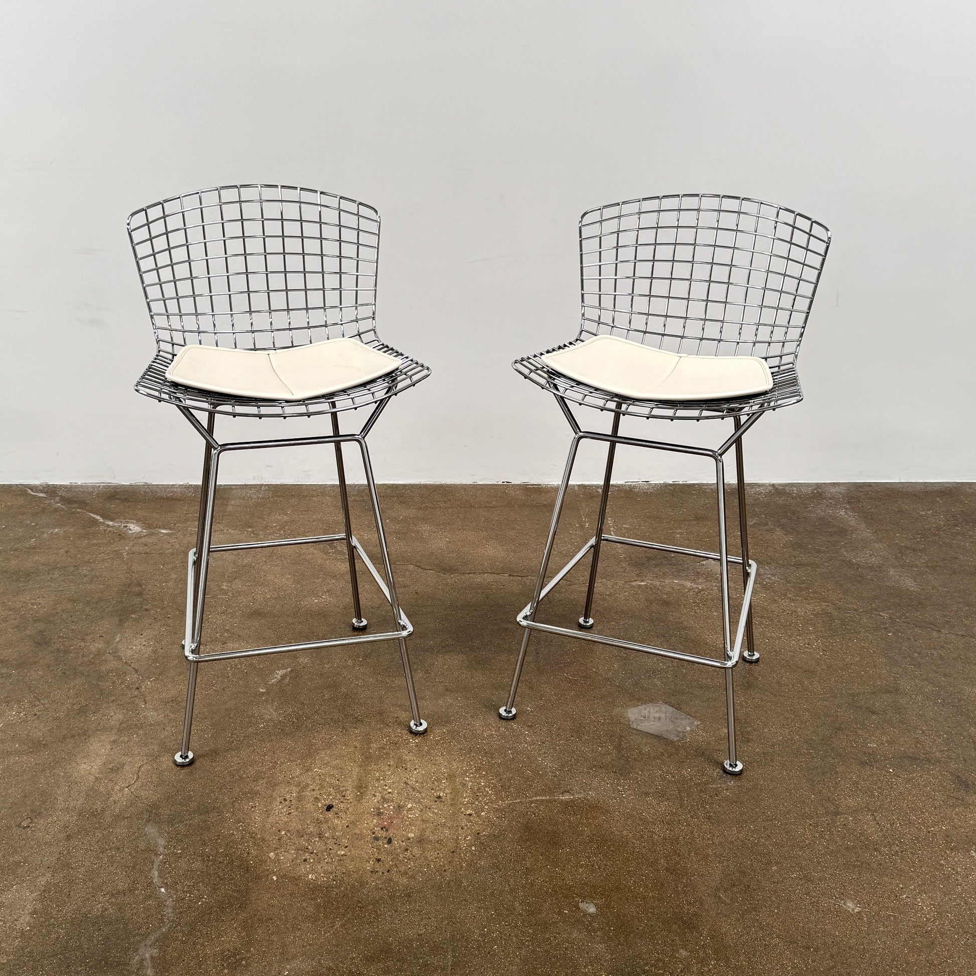 A set of two Knoll Bertoia Counter Stools by Knoll, with light seat cushions, are placed side by side on a brown concrete floor against a plain white wall.