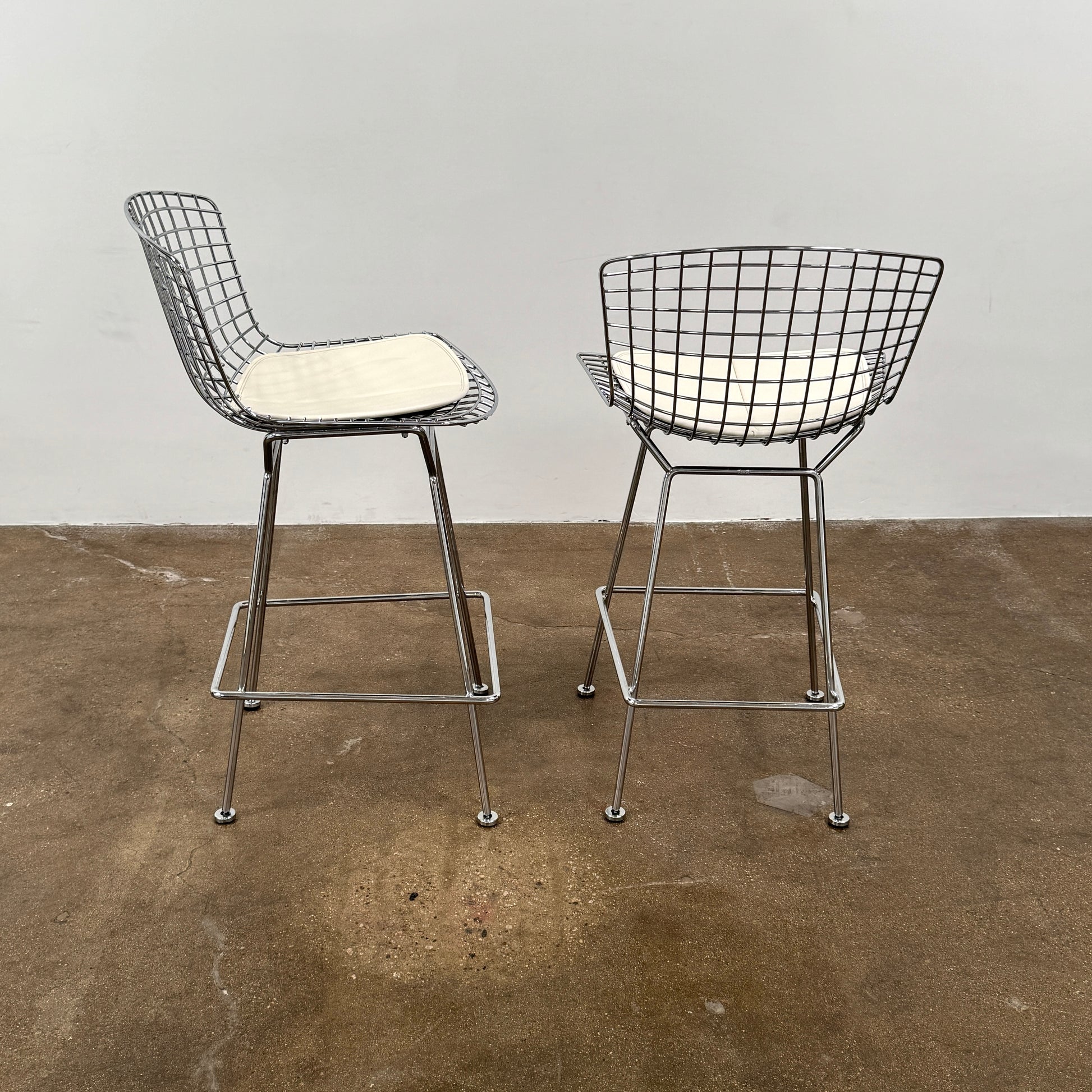 A set of two Knoll Bertoia Counter Stools by Knoll, with light seat cushions, are placed side by side on a brown concrete floor against a plain white wall.