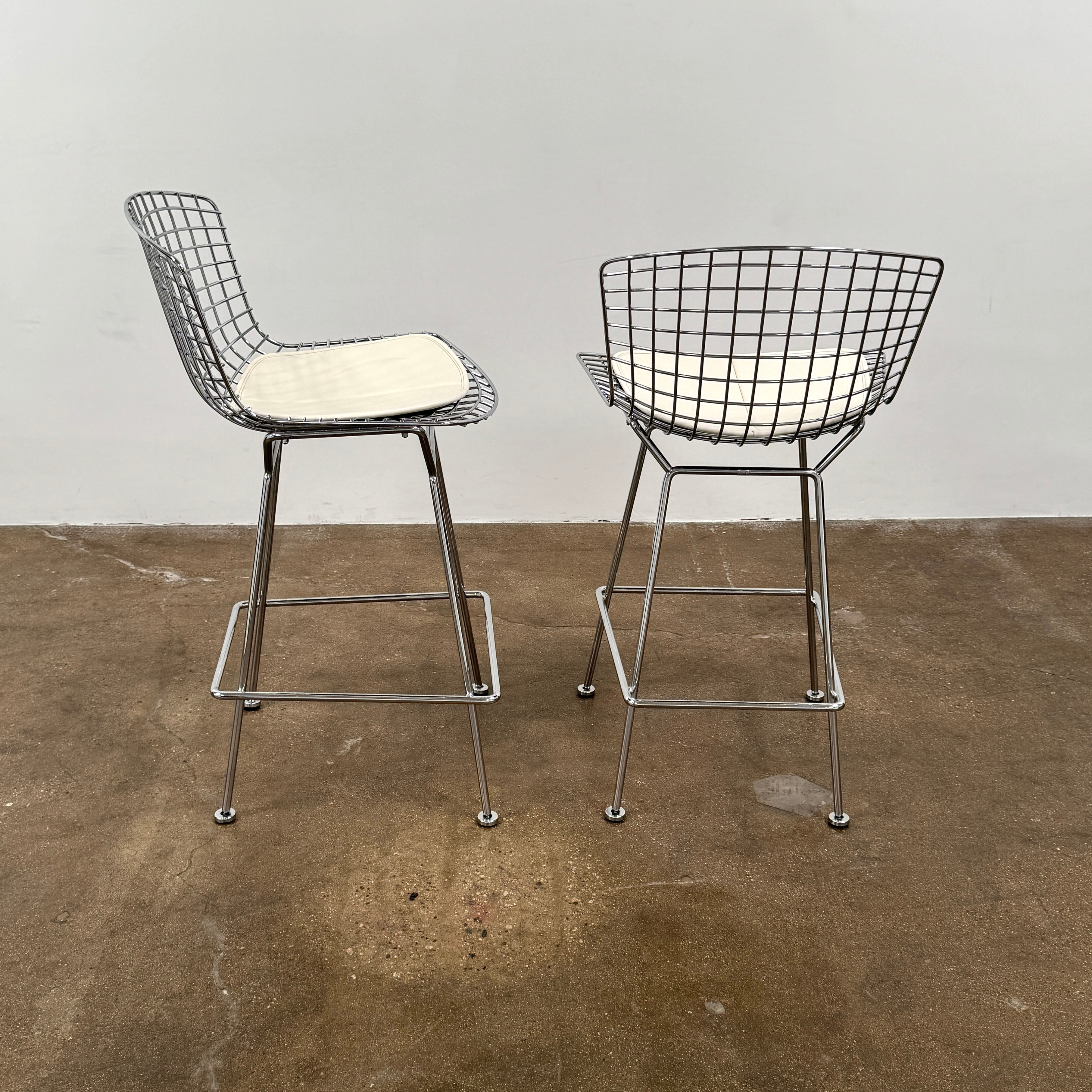 A set of two Knoll Bertoia Counter Stools by Knoll, with light seat cushions, are placed side by side on a brown concrete floor against a plain white wall.
