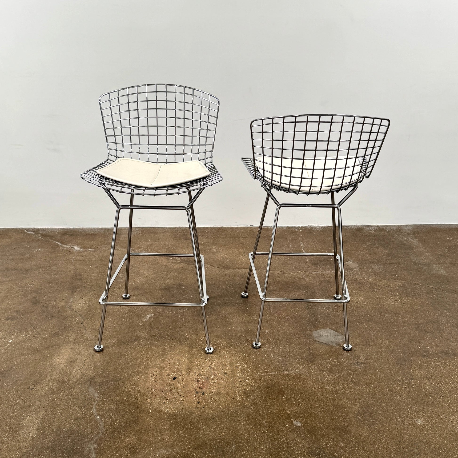 A set of two Knoll Bertoia Counter Stools by Knoll, with light seat cushions, are placed side by side on a brown concrete floor against a plain white wall.