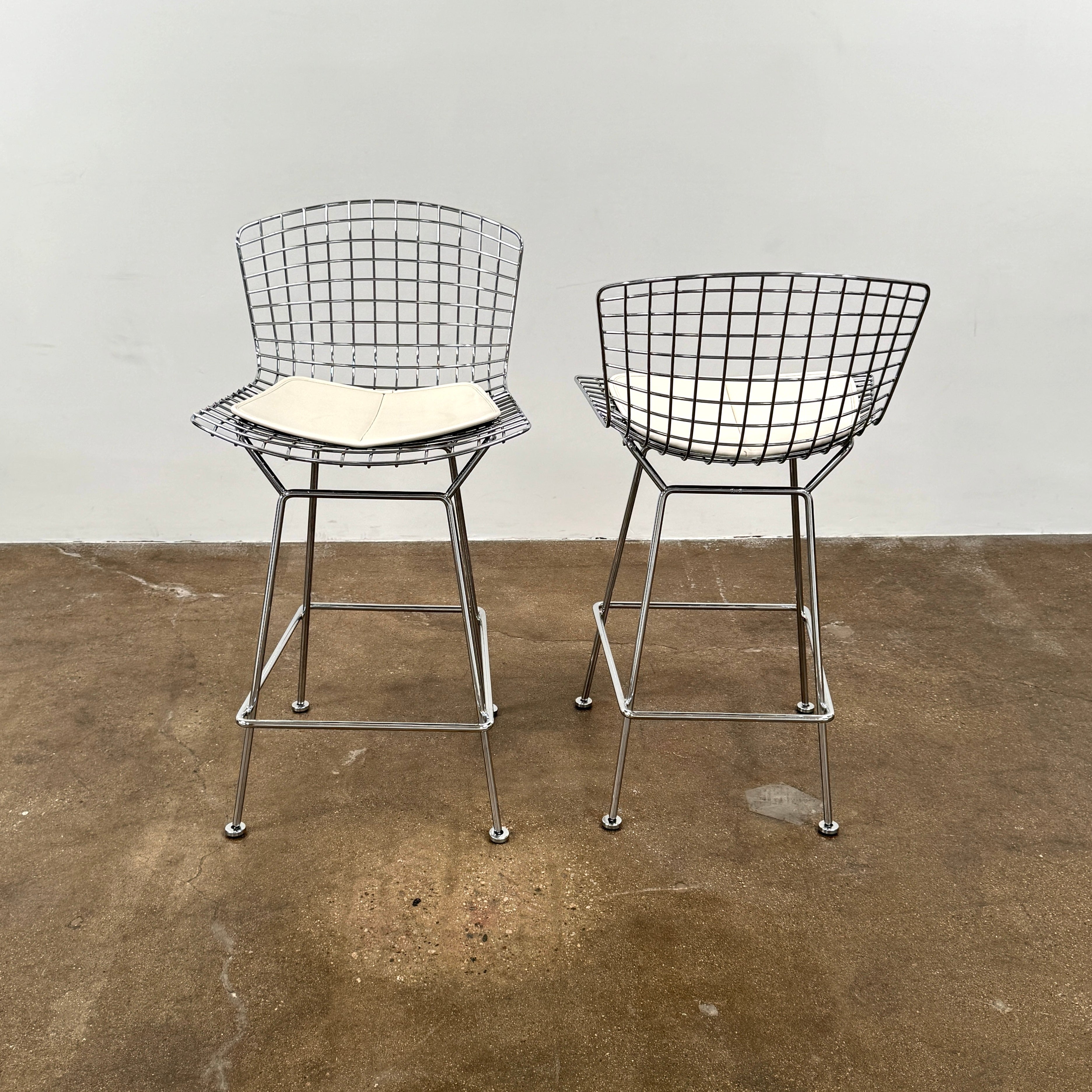 A set of two Knoll Bertoia Counter Stools by Knoll, with light seat cushions, are placed side by side on a brown concrete floor against a plain white wall.