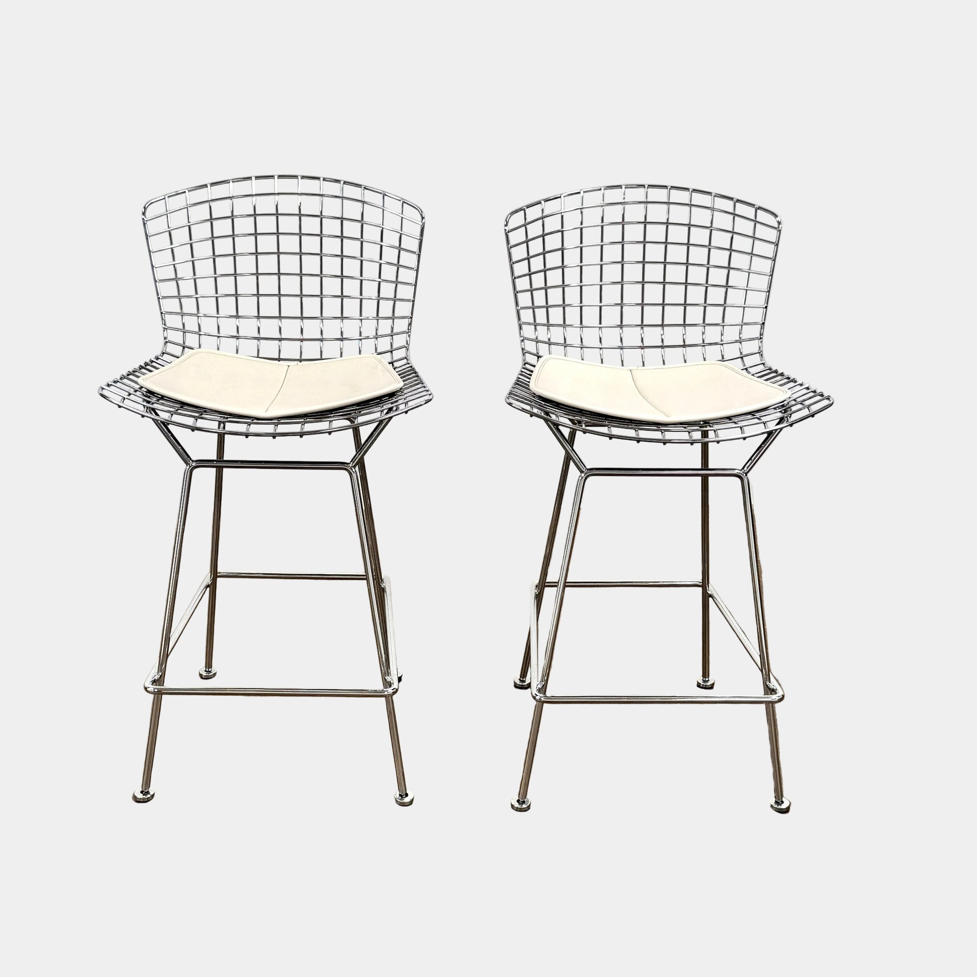 A set of two Knoll Bertoia Counter Stools by Knoll, with light seat cushions, are placed side by side on a brown concrete floor against a plain white wall.