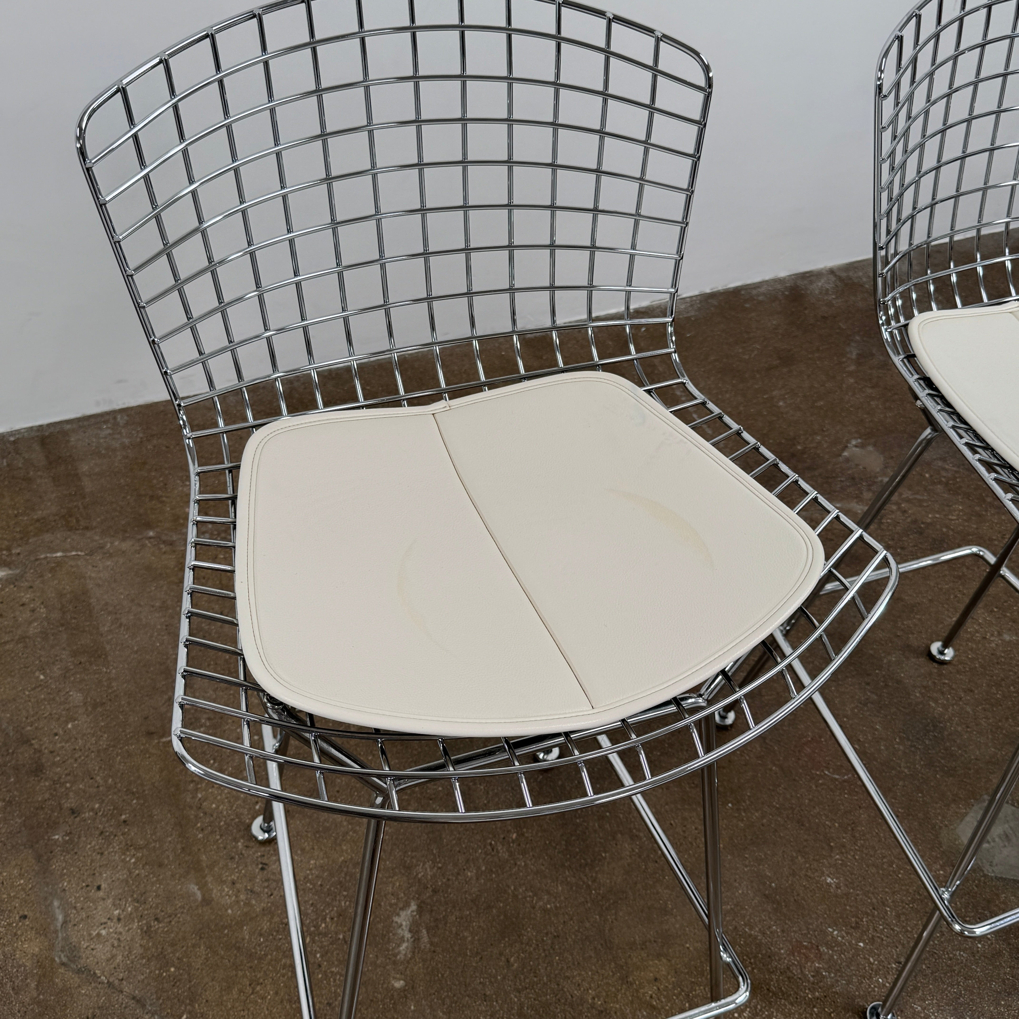 A set of two Knoll Bertoia Counter Stools by Knoll, with light seat cushions, are placed side by side on a brown concrete floor against a plain white wall.