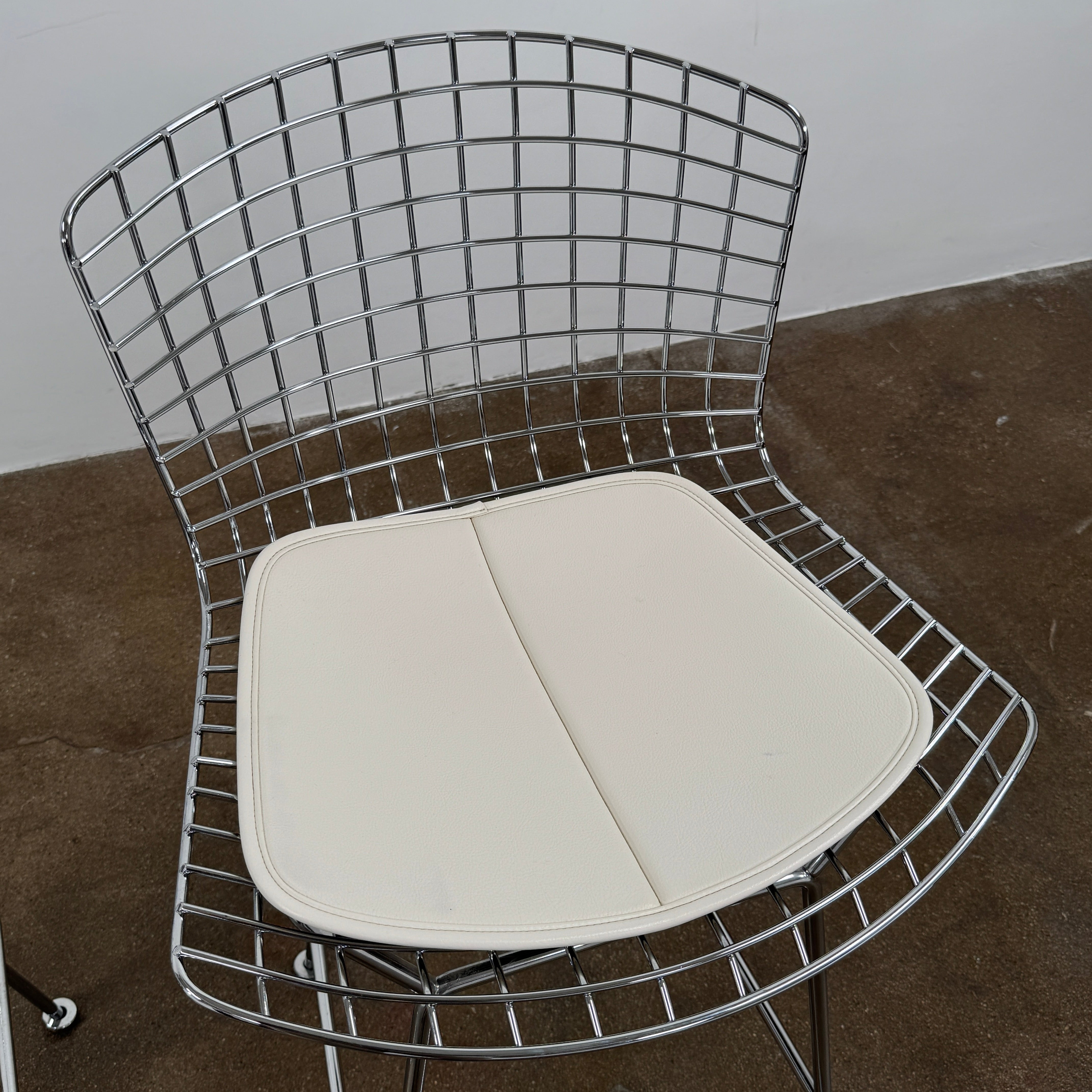 A set of two Knoll Bertoia Counter Stools by Knoll, with light seat cushions, are placed side by side on a brown concrete floor against a plain white wall.