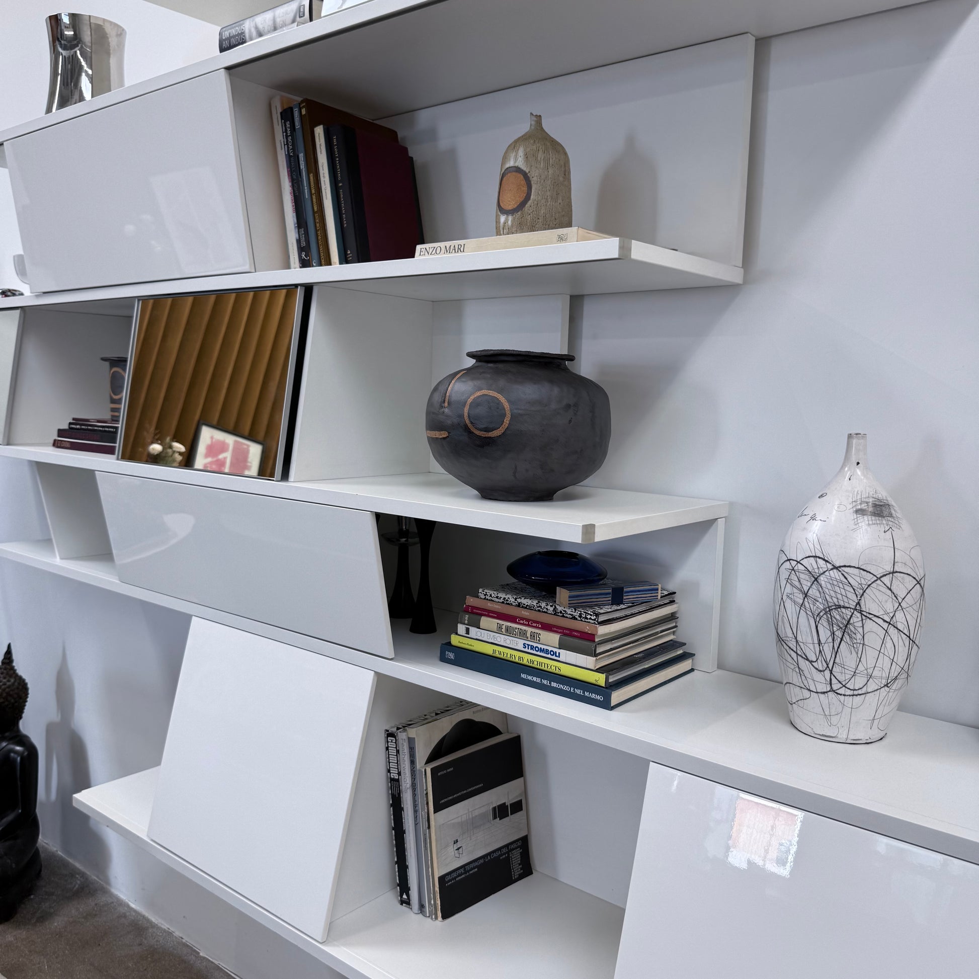 A white, wall-hanging B&B Italia Shift Wall Hanging Shelving System featuring horizontal tiers, enclosed cabinets, and assorted decorative objects and books to create a sculptured bookcase.