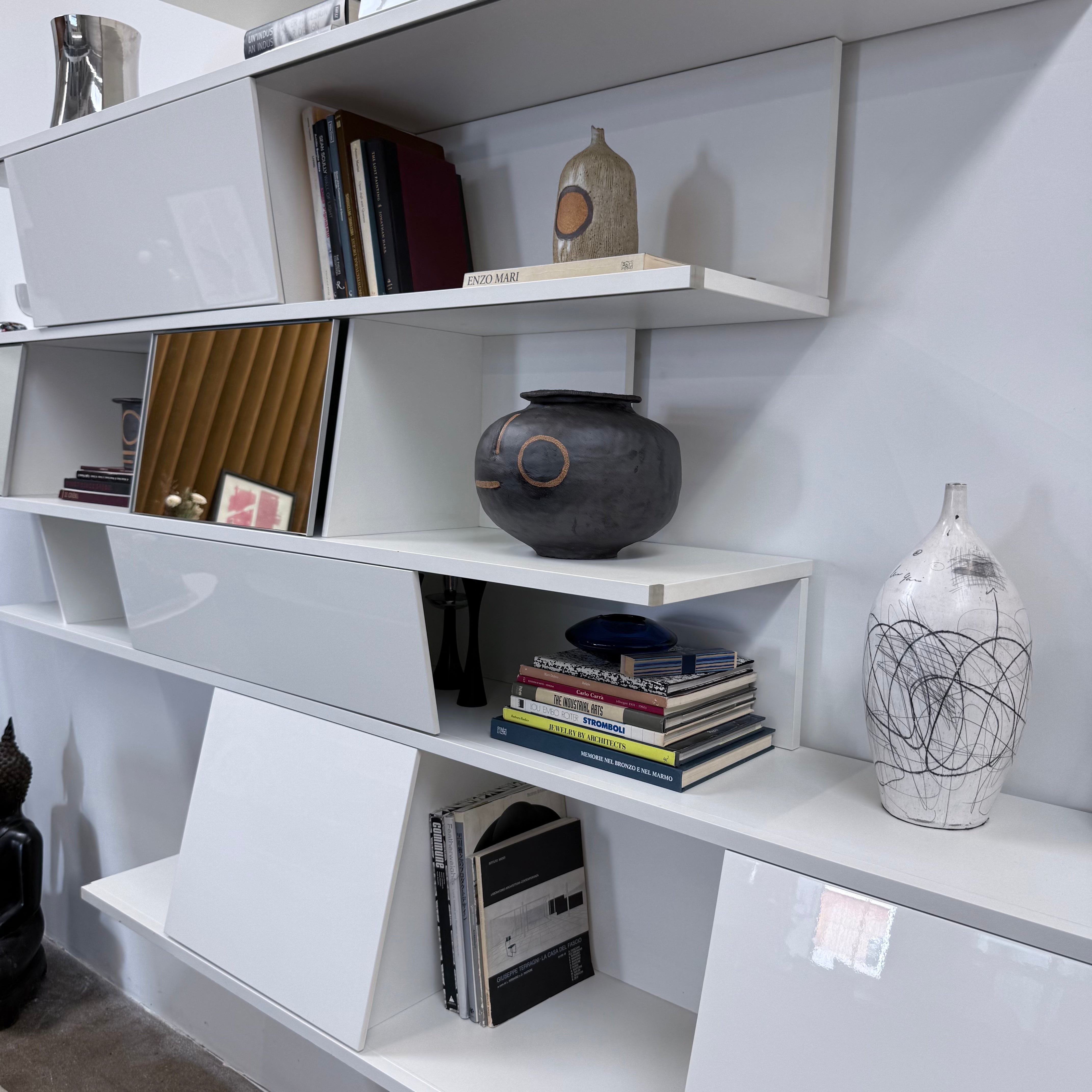 A white, wall-hanging B&B Italia Shift Wall Hanging Shelving System featuring horizontal tiers, enclosed cabinets, and assorted decorative objects and books to create a sculptured bookcase.
