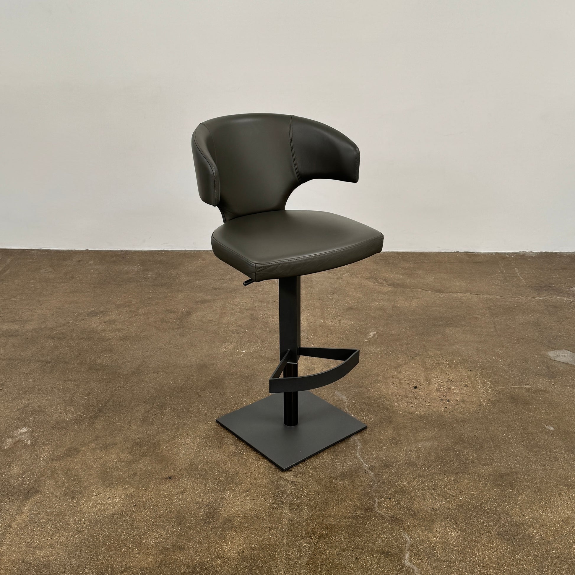 The Cantoni Wing Barstool by Cantoni features a modern black swivel design with a metal base, set on a brown concrete floor against a plain white wall.