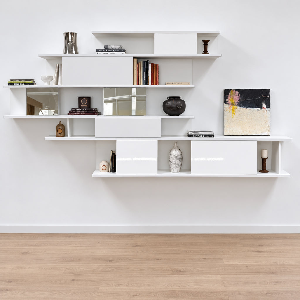 A white, wall-hanging B&B Italia Shift Wall Hanging Shelving System featuring horizontal tiers, enclosed cabinets, and assorted decorative objects and books to create a sculptured bookcase.