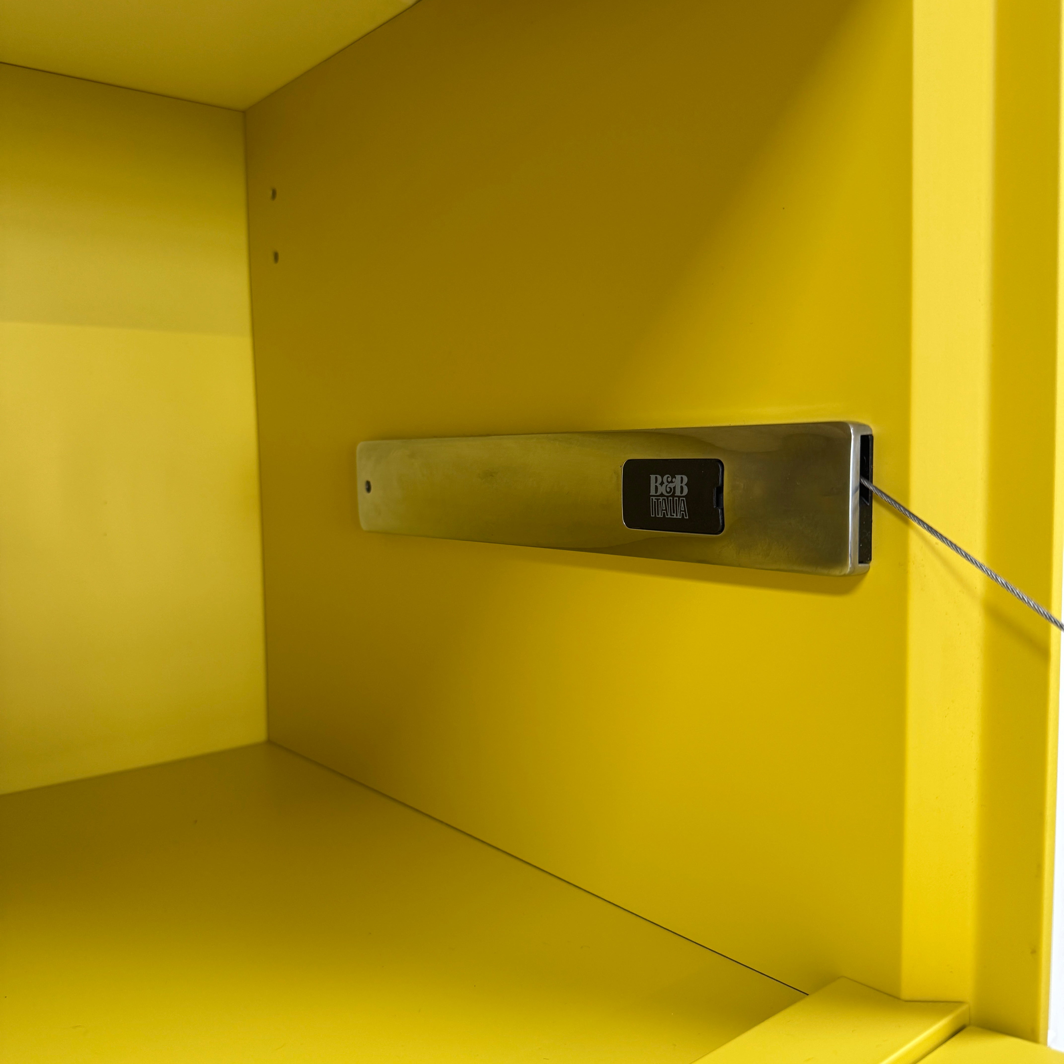Close-up of a yellow cabinet door with a small paint chip on the bottom edge; black bowls and books are seen on a white shelf of the B&B Italia Flat C Wall Unit With Storage by B&B Italia, combining style and function.