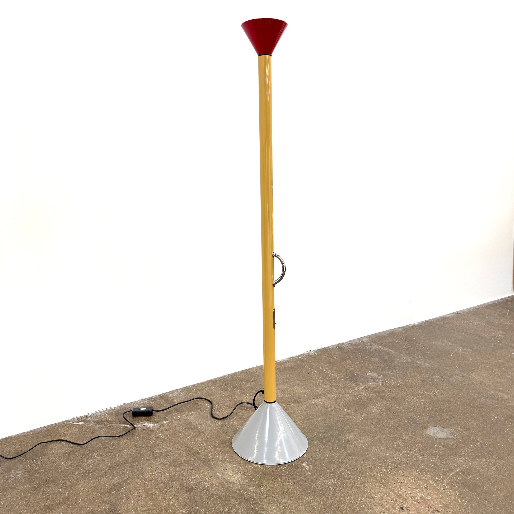 A bold yellow Artemide Callimaco Floor Lamp by Artimide, with a red cone top, silver conical base, and black cord, stands on a concrete floor against a white wall—a striking example of Radical Design by Ettore Sottsass.