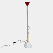 A bold yellow Artemide Callimaco Floor Lamp by Artimide, with a red cone top, silver conical base, and black cord, stands on a concrete floor against a white wall—a striking example of Radical Design by Ettore Sottsass.