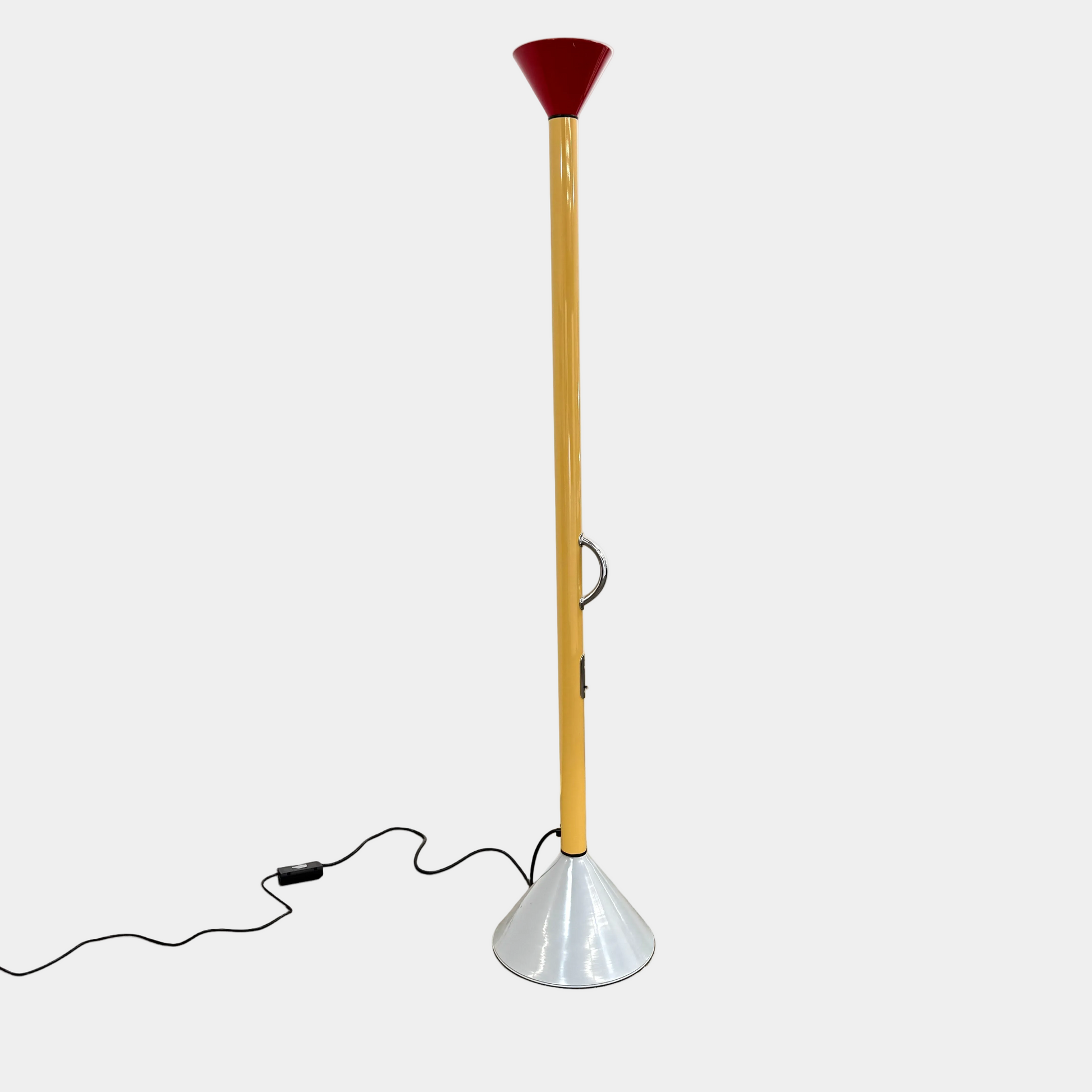 A bold yellow Artemide Callimaco Floor Lamp by Artimide, with a red cone top, silver conical base, and black cord, stands on a concrete floor against a white wall—a striking example of Radical Design by Ettore Sottsass.