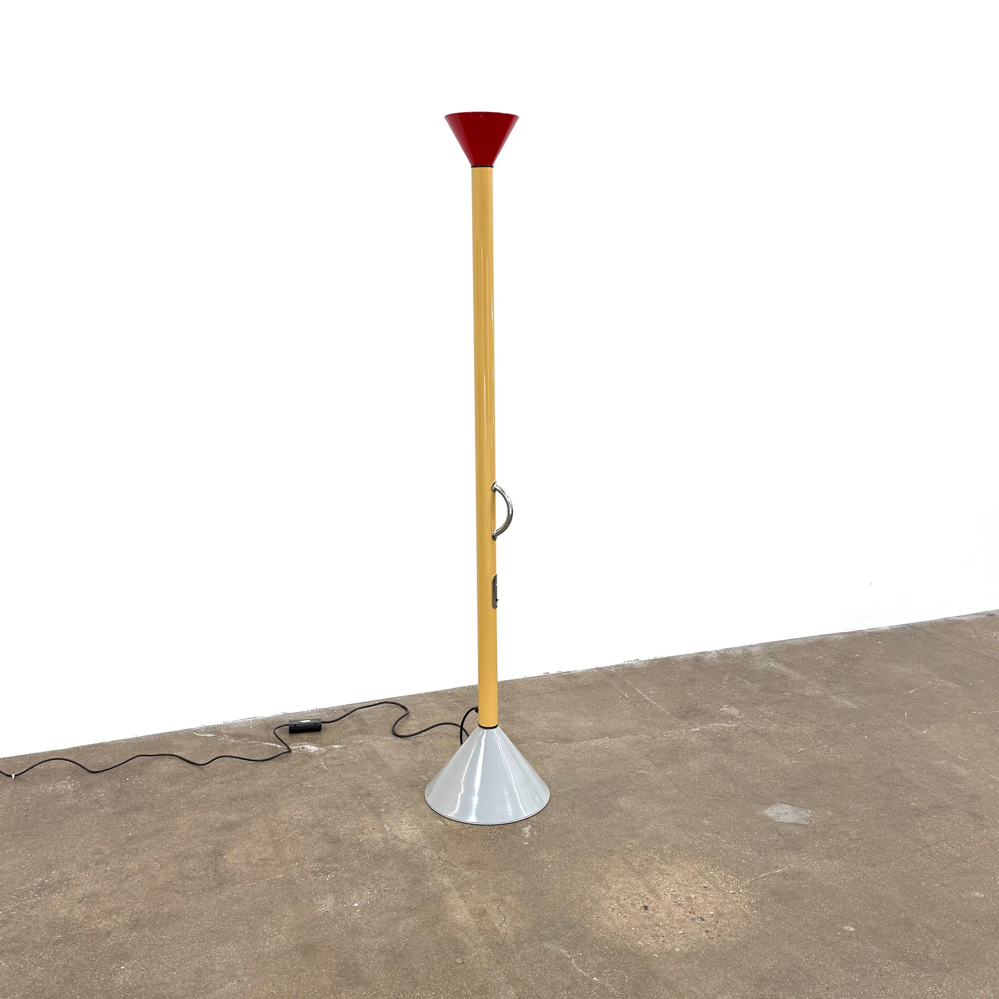 A bold yellow Artemide Callimaco Floor Lamp by Artimide, with a red cone top, silver conical base, and black cord, stands on a concrete floor against a white wall—a striking example of Radical Design by Ettore Sottsass.