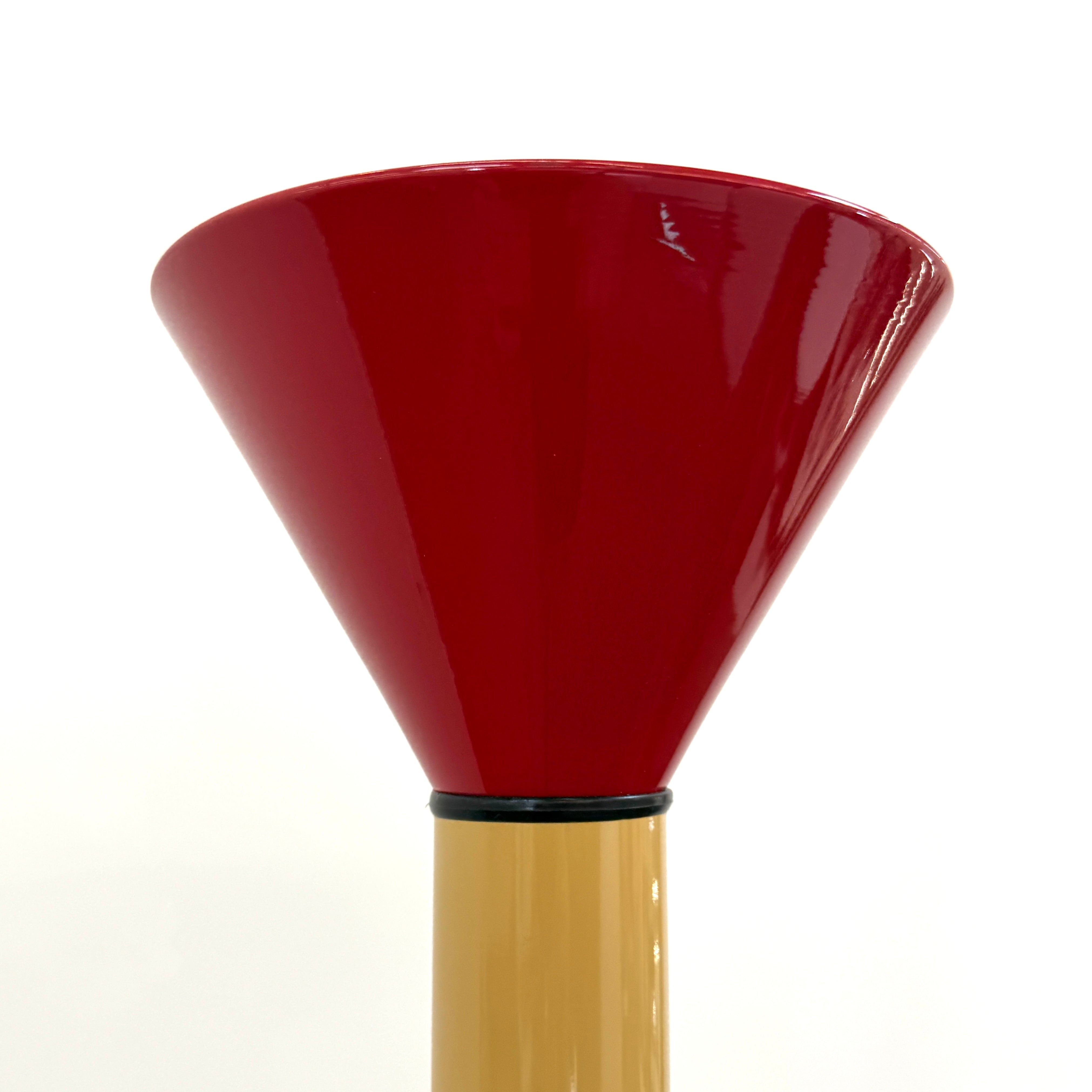 A bold yellow Artemide Callimaco Floor Lamp by Artimide, with a red cone top, silver conical base, and black cord, stands on a concrete floor against a white wall—a striking example of Radical Design by Ettore Sottsass.