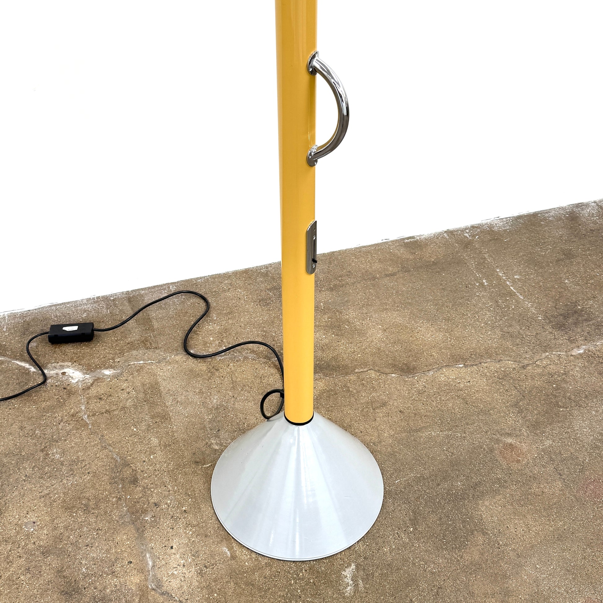 A bold yellow Artemide Callimaco Floor Lamp by Artimide, with a red cone top, silver conical base, and black cord, stands on a concrete floor against a white wall—a striking example of Radical Design by Ettore Sottsass.