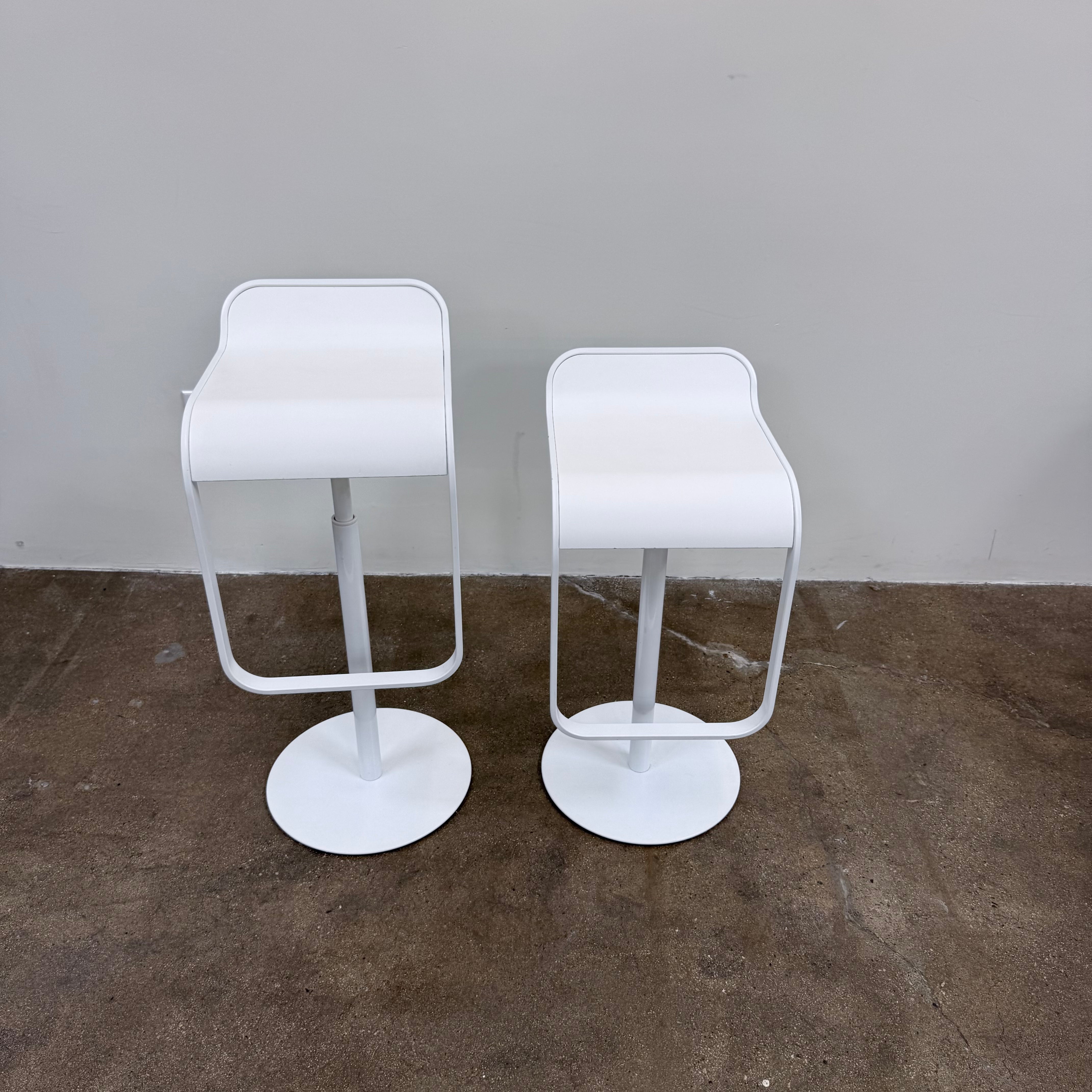 Two white LEM Piston Stools by La Palma featuring pedestal bases and rectangular footrests.