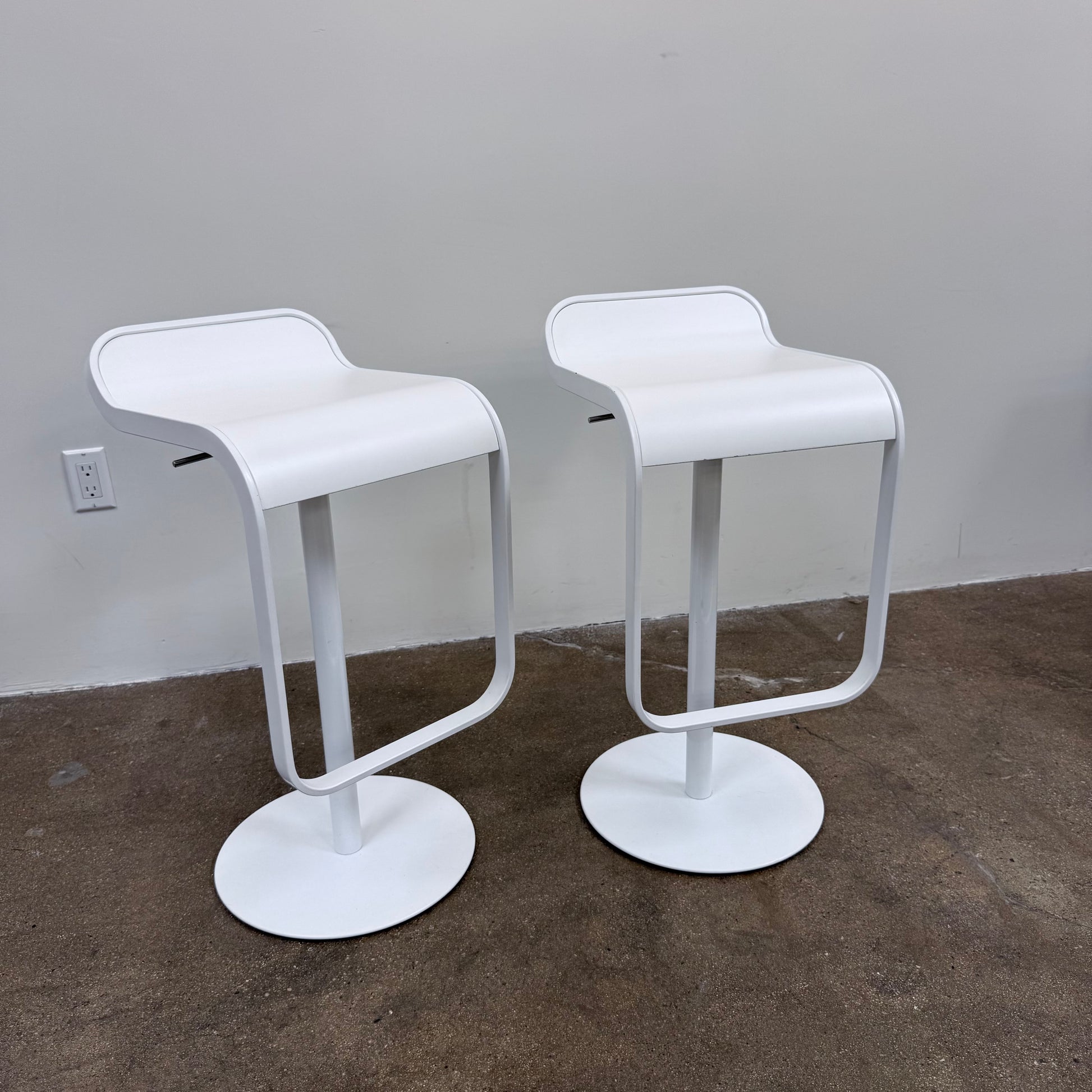 Two white LEM Piston Stools by La Palma featuring pedestal bases and rectangular footrests.