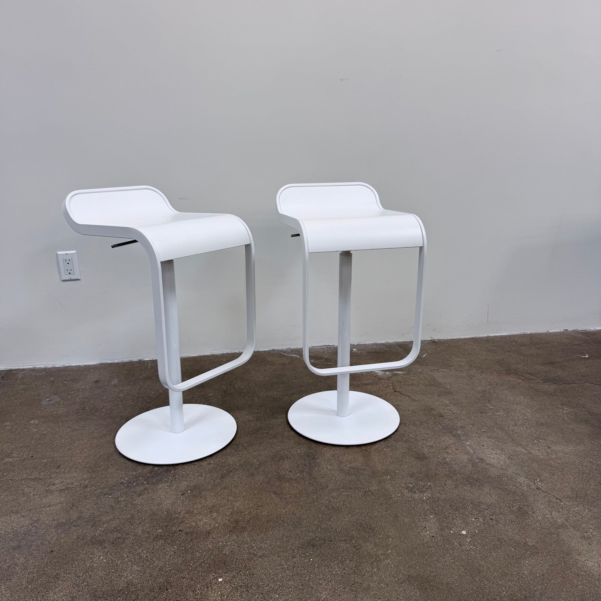 Two white LEM Piston Stools by La Palma featuring pedestal bases and rectangular footrests.