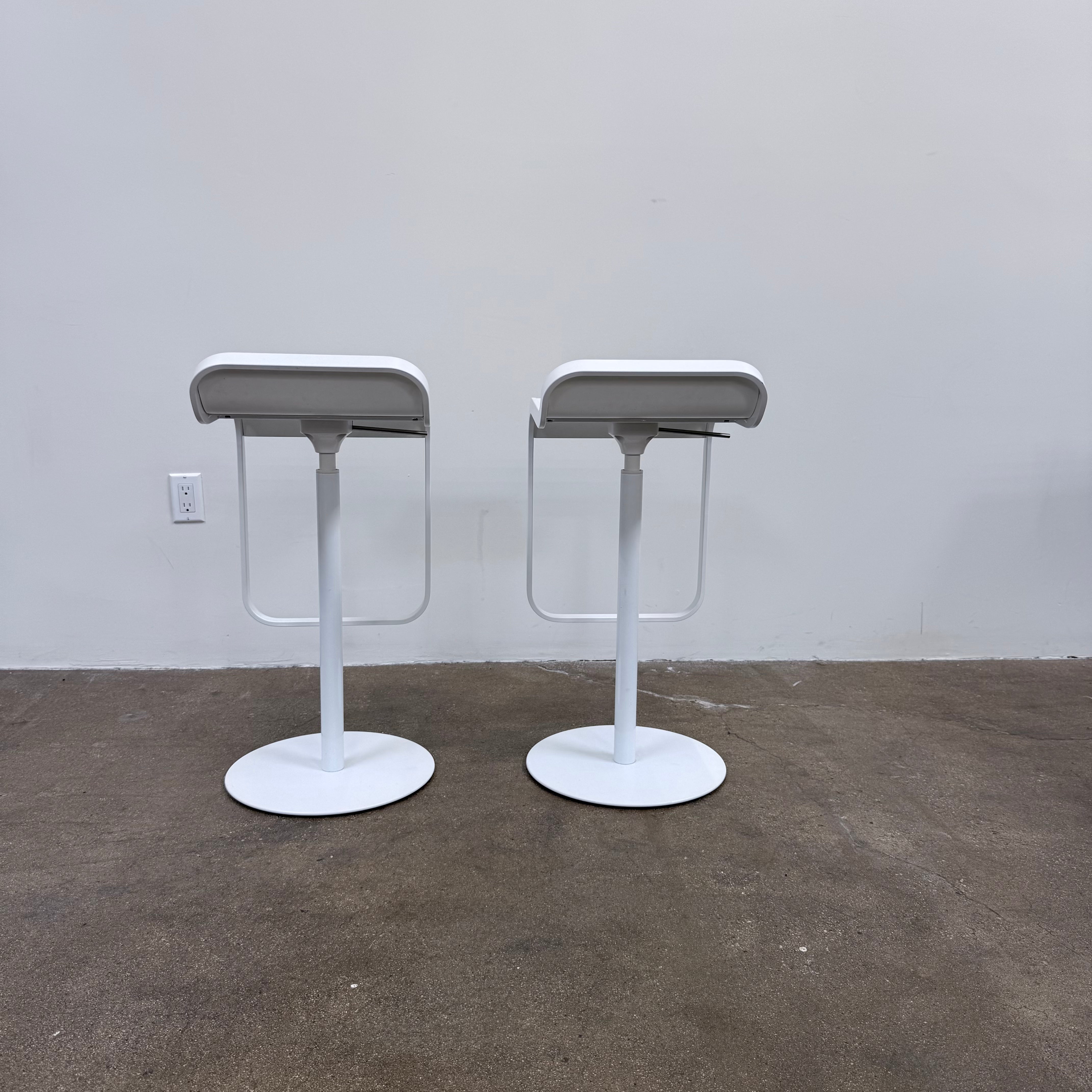 Two white LEM Piston Stools by La Palma featuring pedestal bases and rectangular footrests.