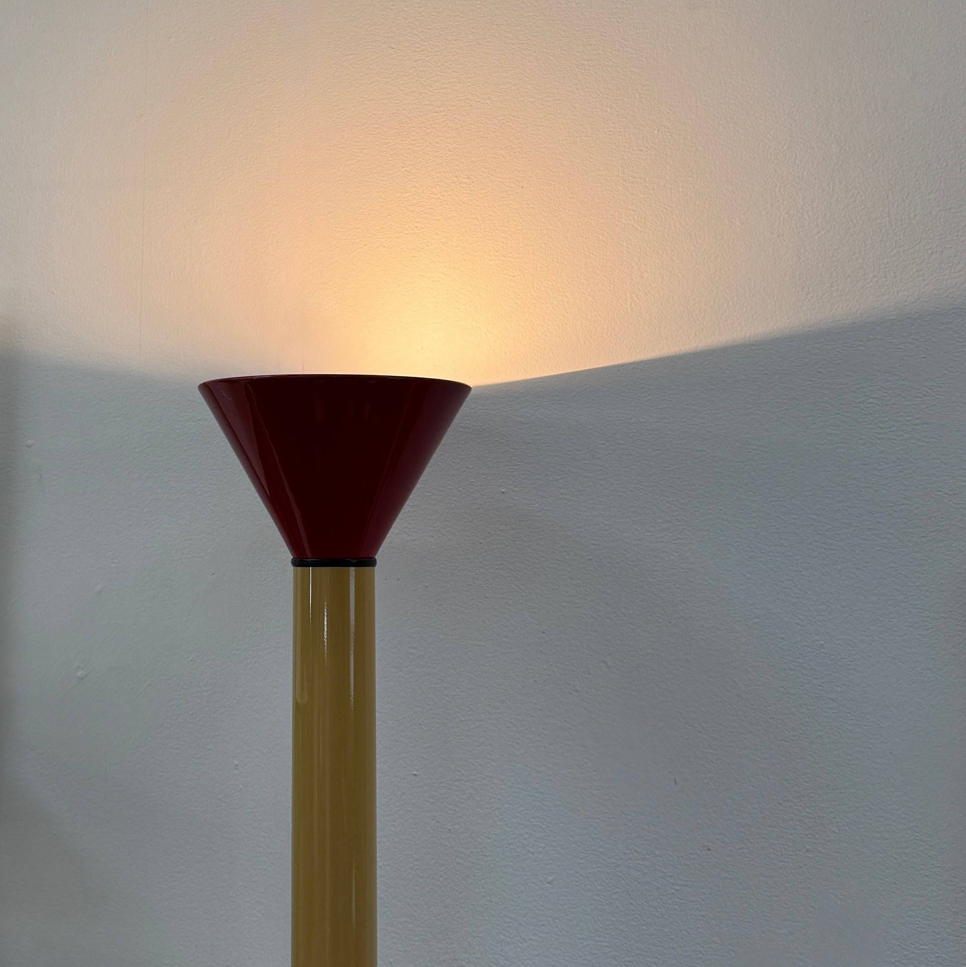 A bold yellow Artemide Callimaco Floor Lamp by Artimide, with a red cone top, silver conical base, and black cord, stands on a concrete floor against a white wall—a striking example of Radical Design by Ettore Sottsass.