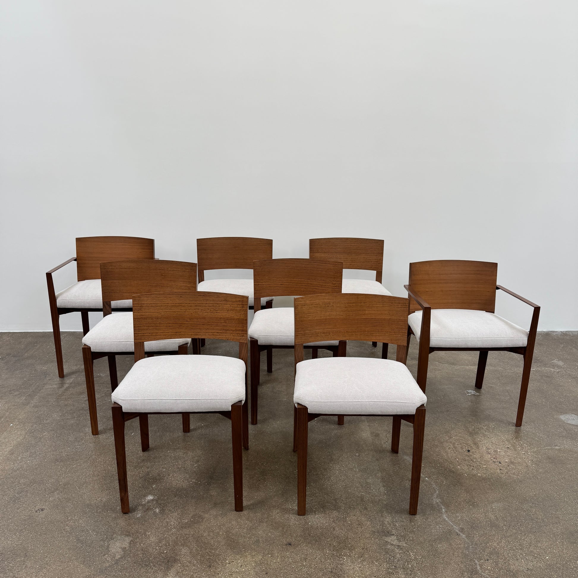A set of 8 Porro Lario dining chairs by Porro, featuring minimalist armrests and light-cushioned seats, sits on a concrete floor against a plain white wall, reflecting refined contemporary style.