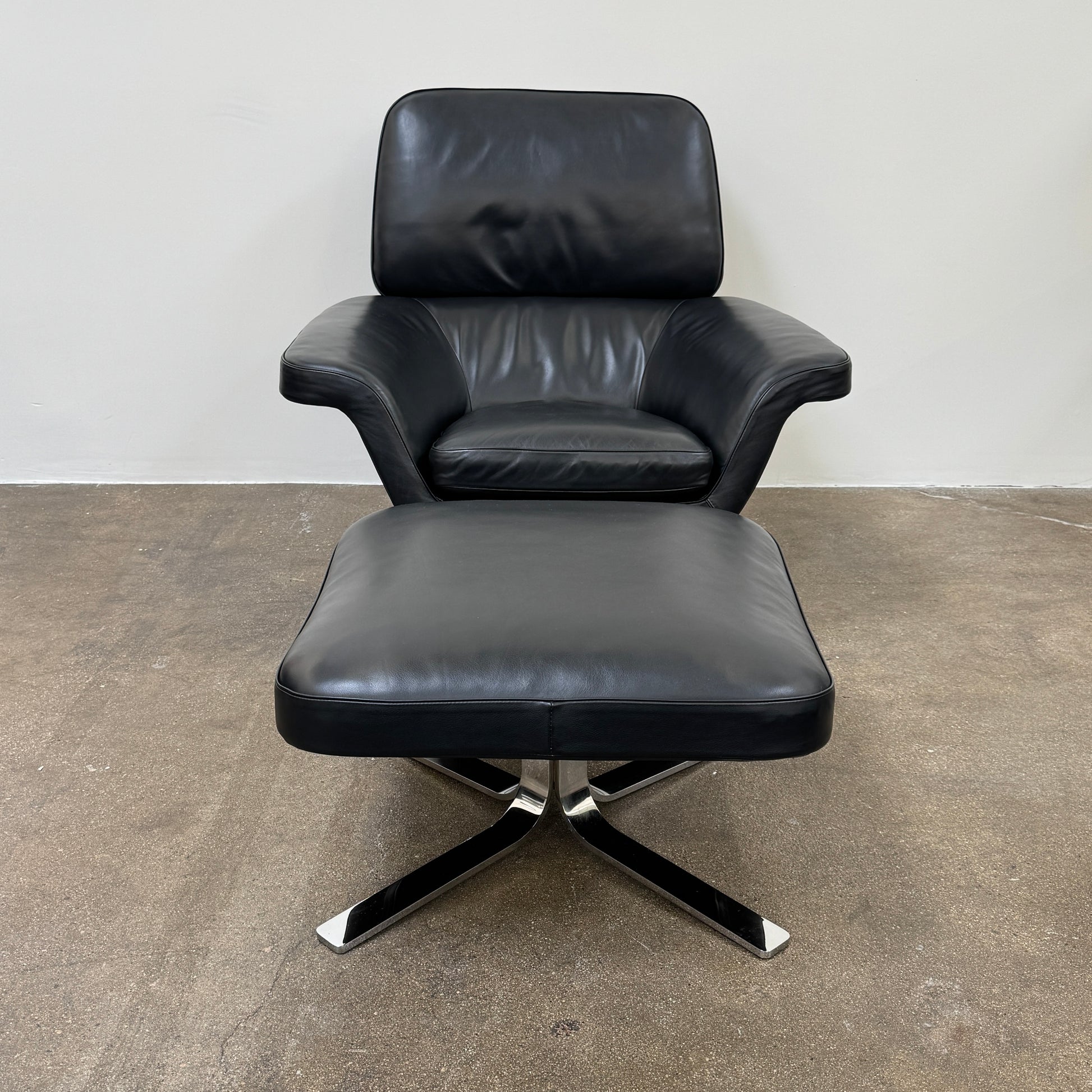 A Minotti Blake Lounge Chair & Ottoman by Minotti in black leather with chrome swivel bases, positioned on a bare concrete floor against a plain light-colored wall.