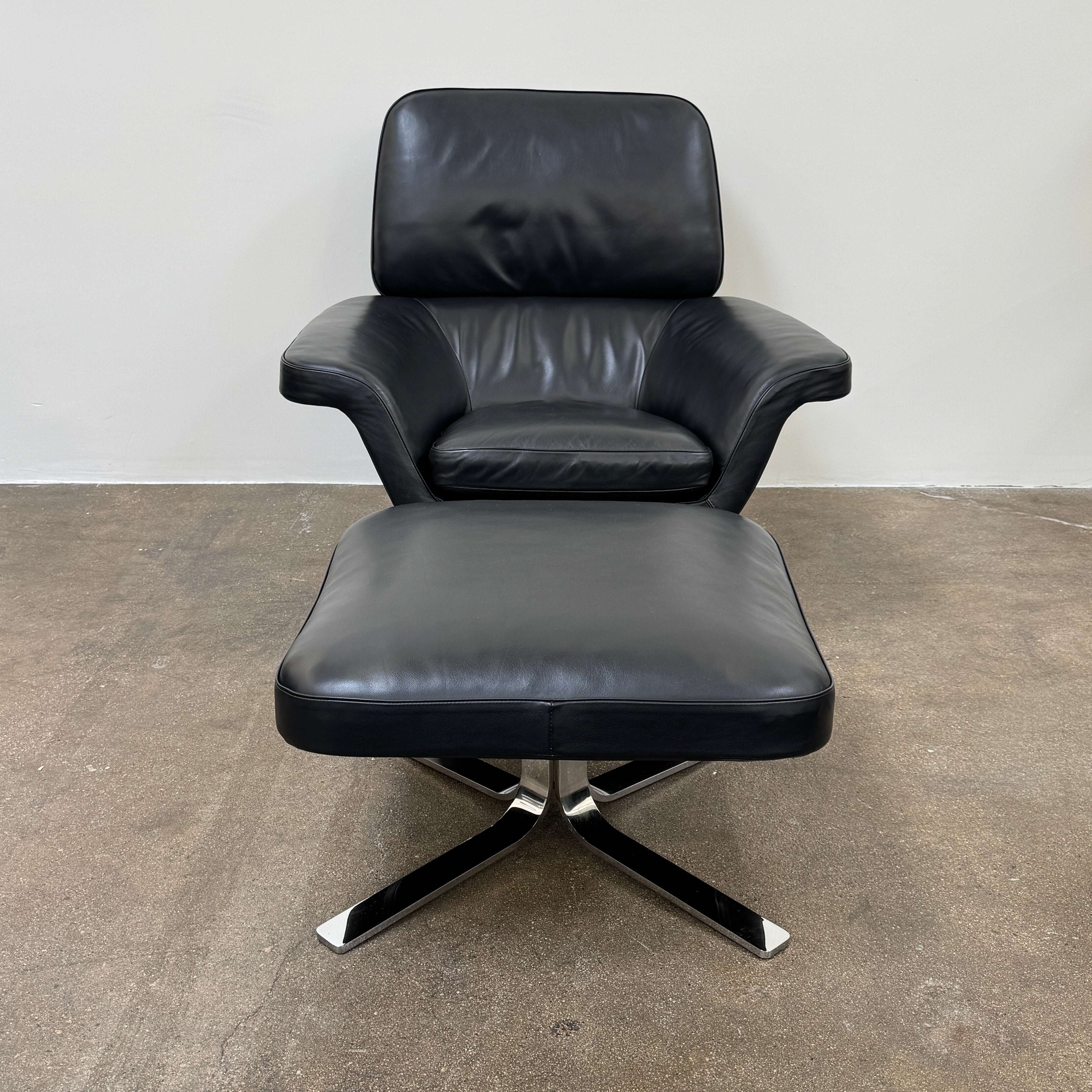 A Minotti Blake Lounge Chair & Ottoman by Minotti in black leather with chrome swivel bases, positioned on a bare concrete floor against a plain light-colored wall.