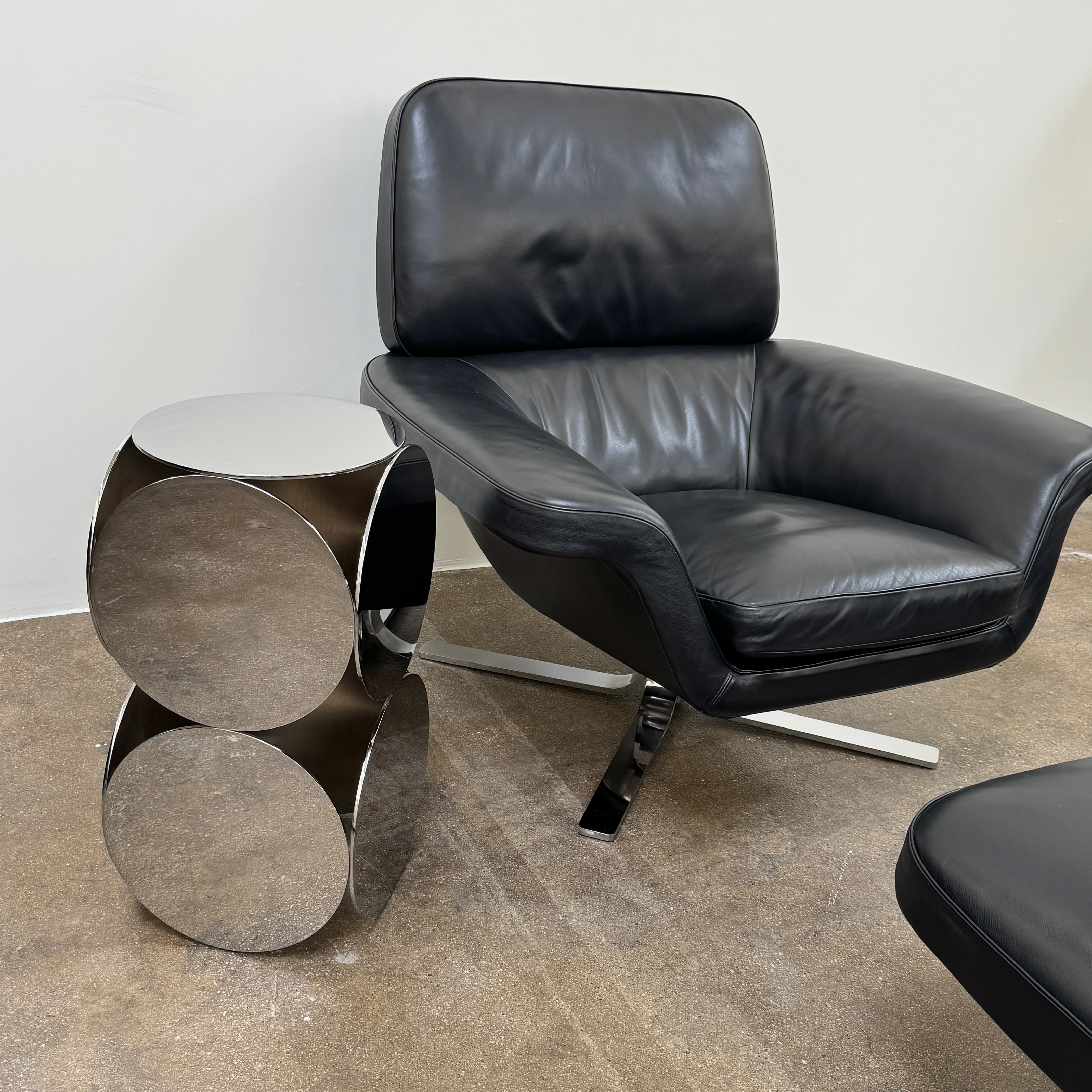 A Minotti Blake Lounge Chair & Ottoman by Minotti in black leather with chrome swivel bases, positioned on a bare concrete floor against a plain light-colored wall.