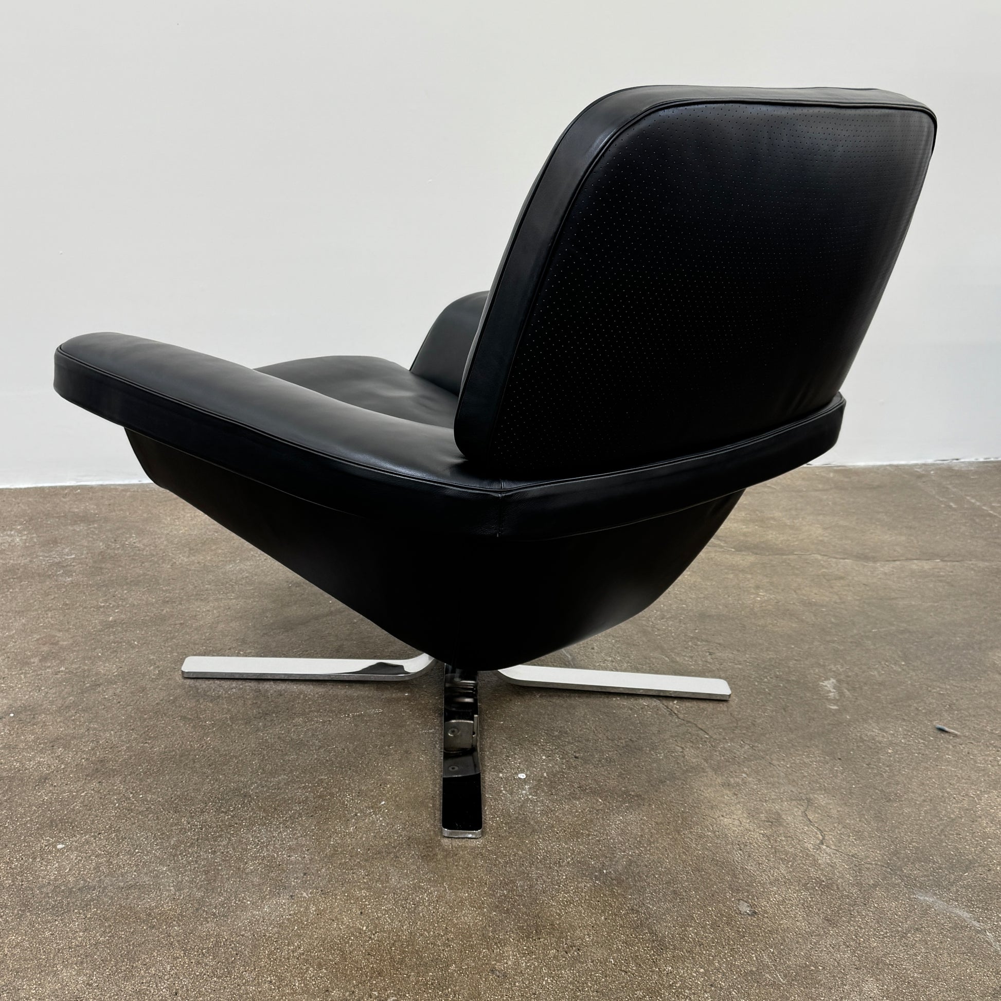 A Minotti Blake Lounge Chair & Ottoman by Minotti in black leather with chrome swivel bases, positioned on a bare concrete floor against a plain light-colored wall.