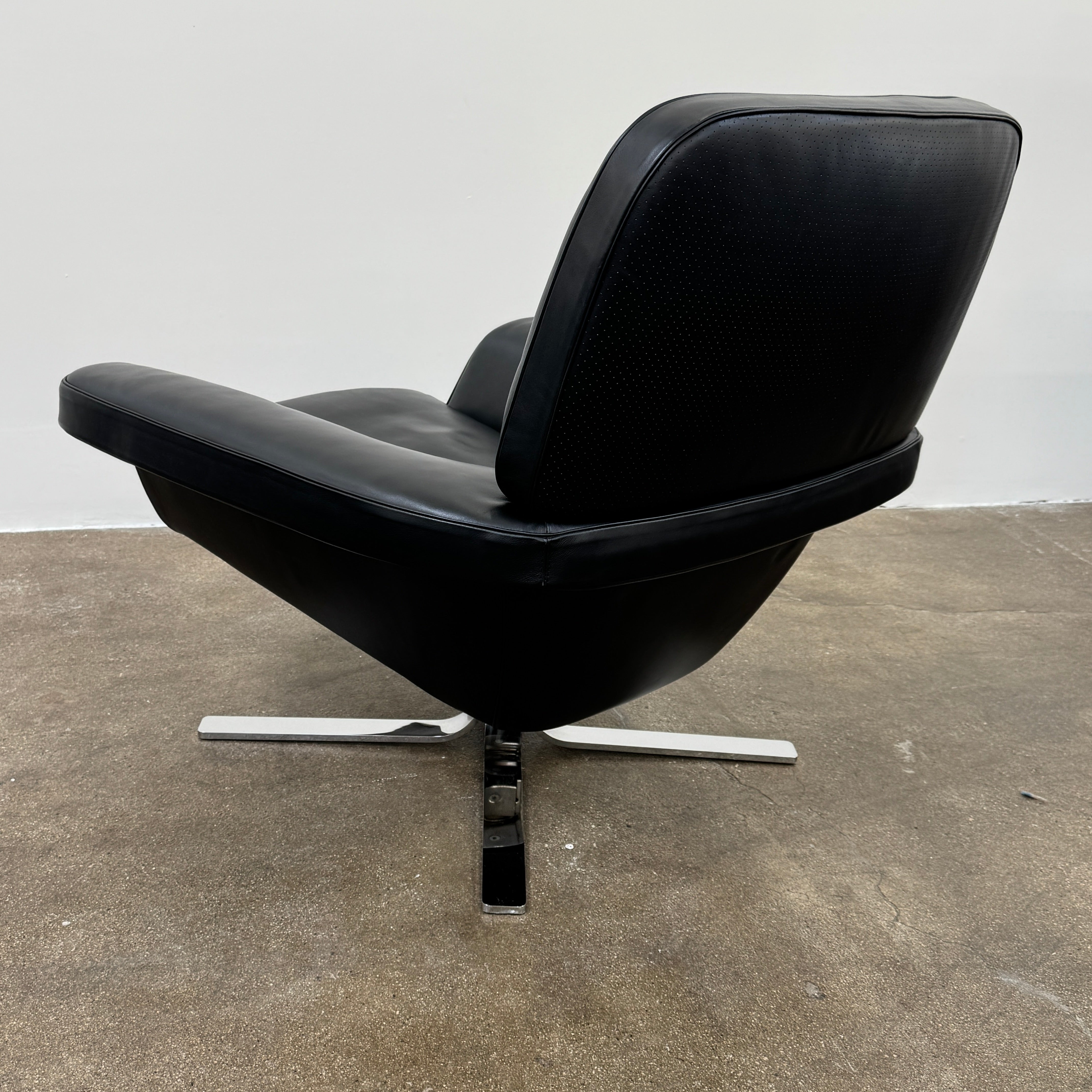 A Minotti Blake Lounge Chair & Ottoman by Minotti in black leather with chrome swivel bases, positioned on a bare concrete floor against a plain light-colored wall.