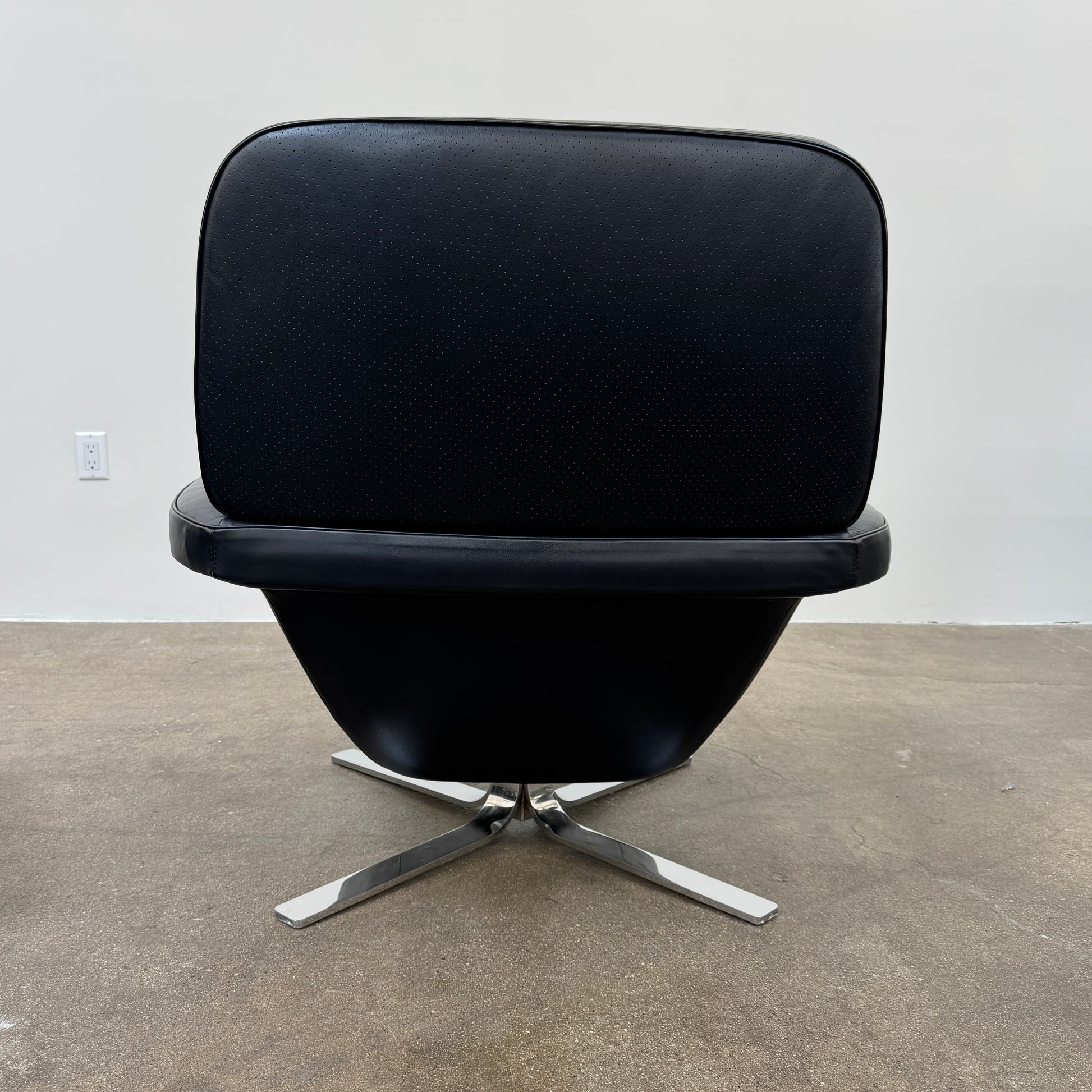 A Minotti Blake Lounge Chair & Ottoman by Minotti in black leather with chrome swivel bases, positioned on a bare concrete floor against a plain light-colored wall.