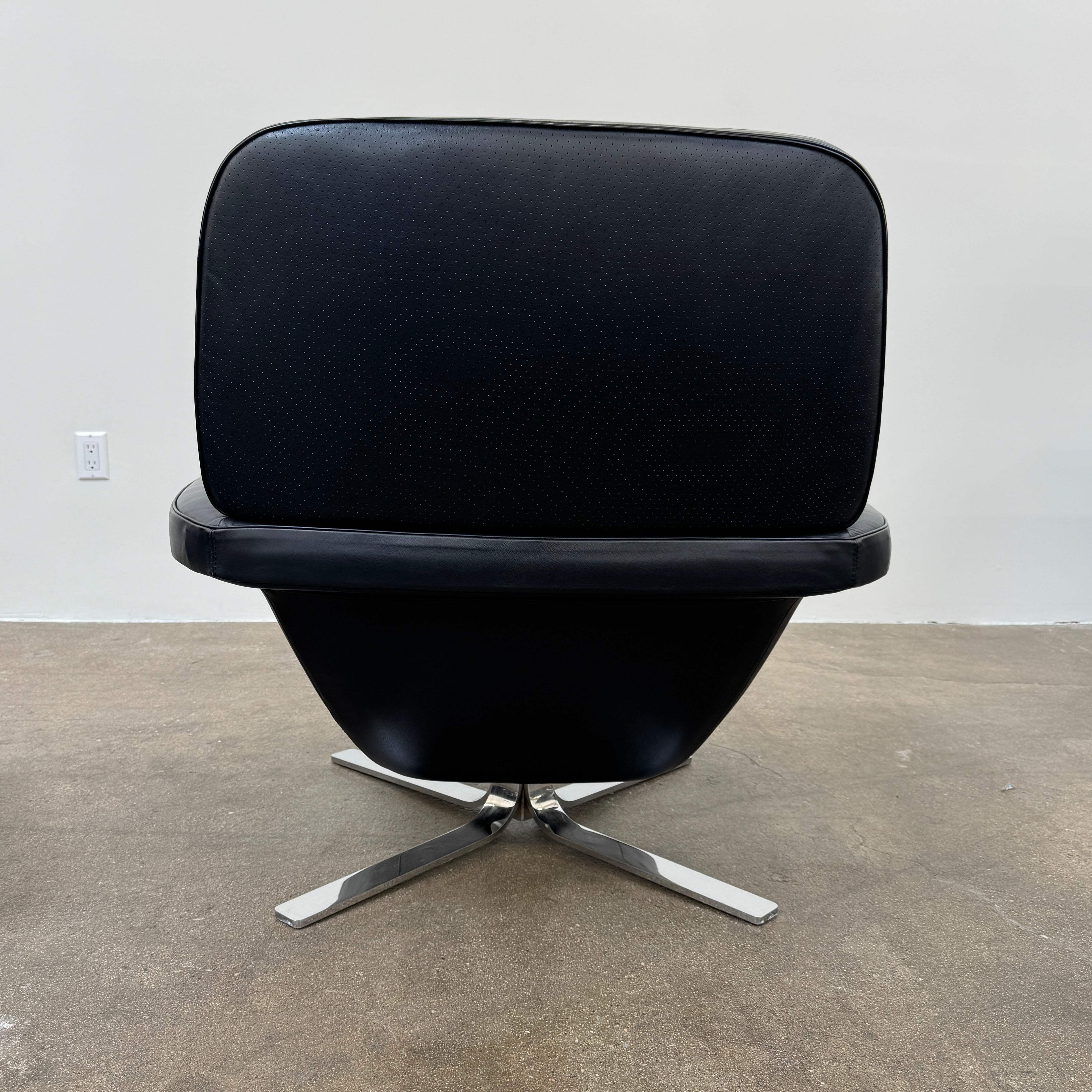 A Minotti Blake Lounge Chair & Ottoman by Minotti in black leather with chrome swivel bases, positioned on a bare concrete floor against a plain light-colored wall.